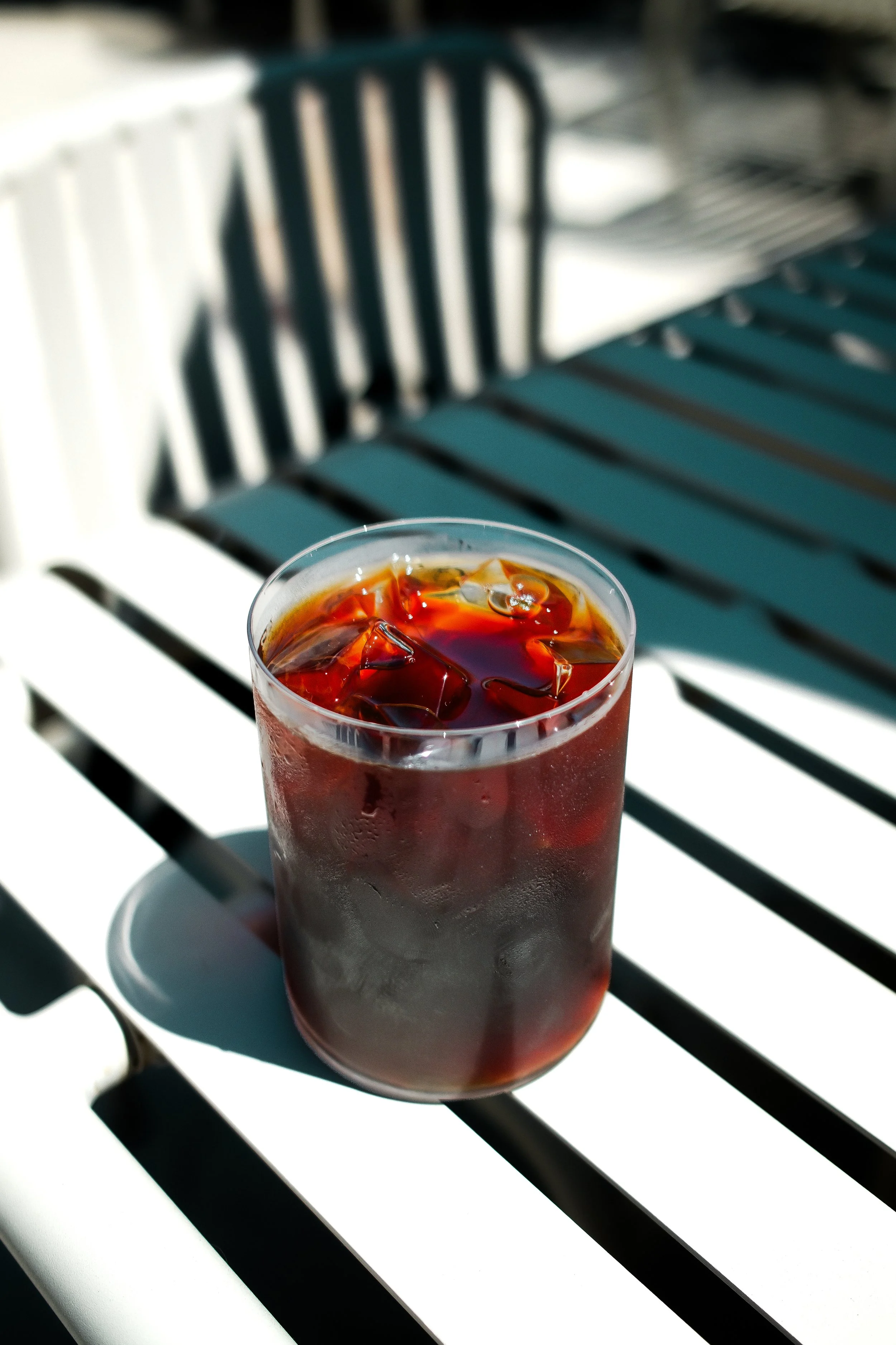 A glass of iced cola on a white saucer with a black straw, placed on a white table with black slats, in bright sunlight.