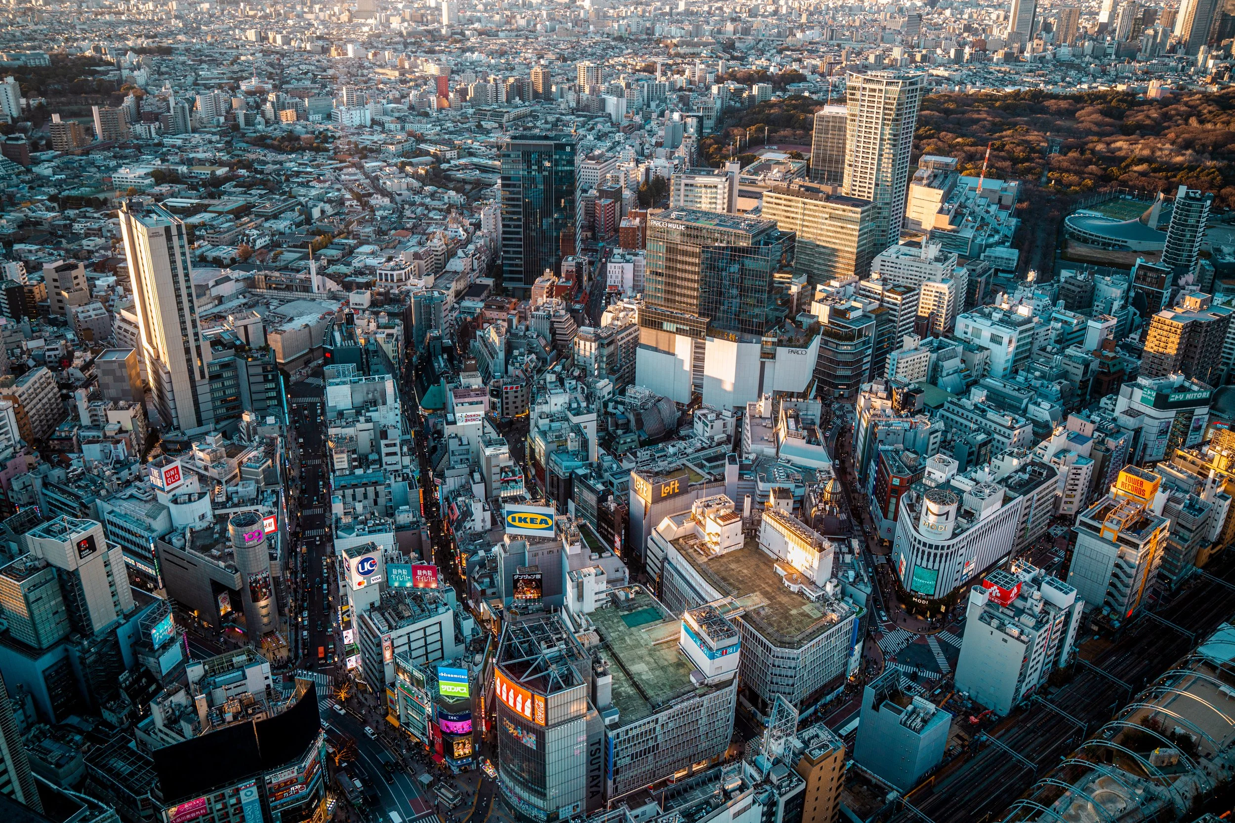 Aerial view of a densely packed cityscape with numerous tall buildings, billboards, and streets, likely in downtown Tokyo during daylight hours.