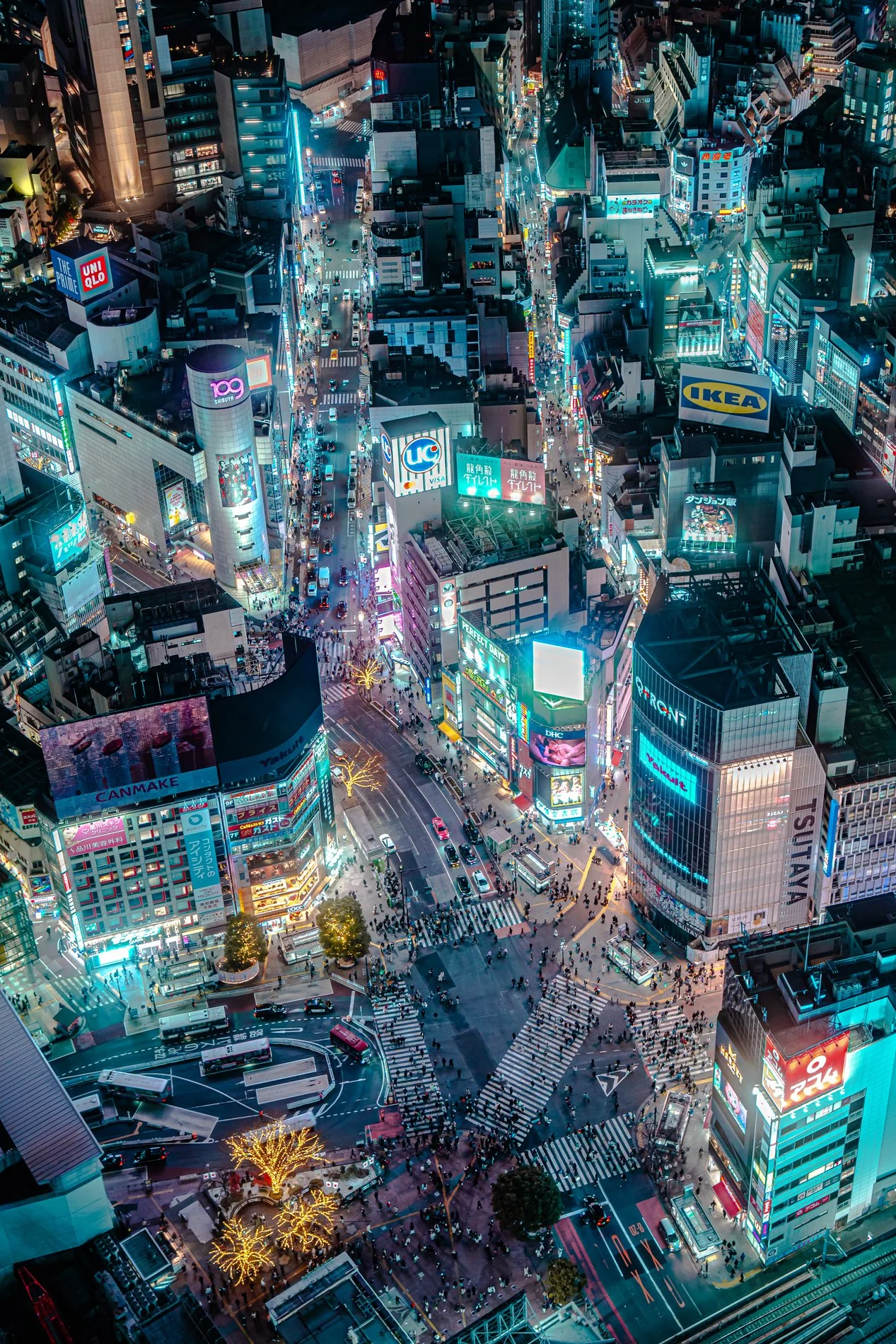 Aerial view of a busy city intersection at night with bright neon signs, tall skyscrapers, and crowded sidewalks.