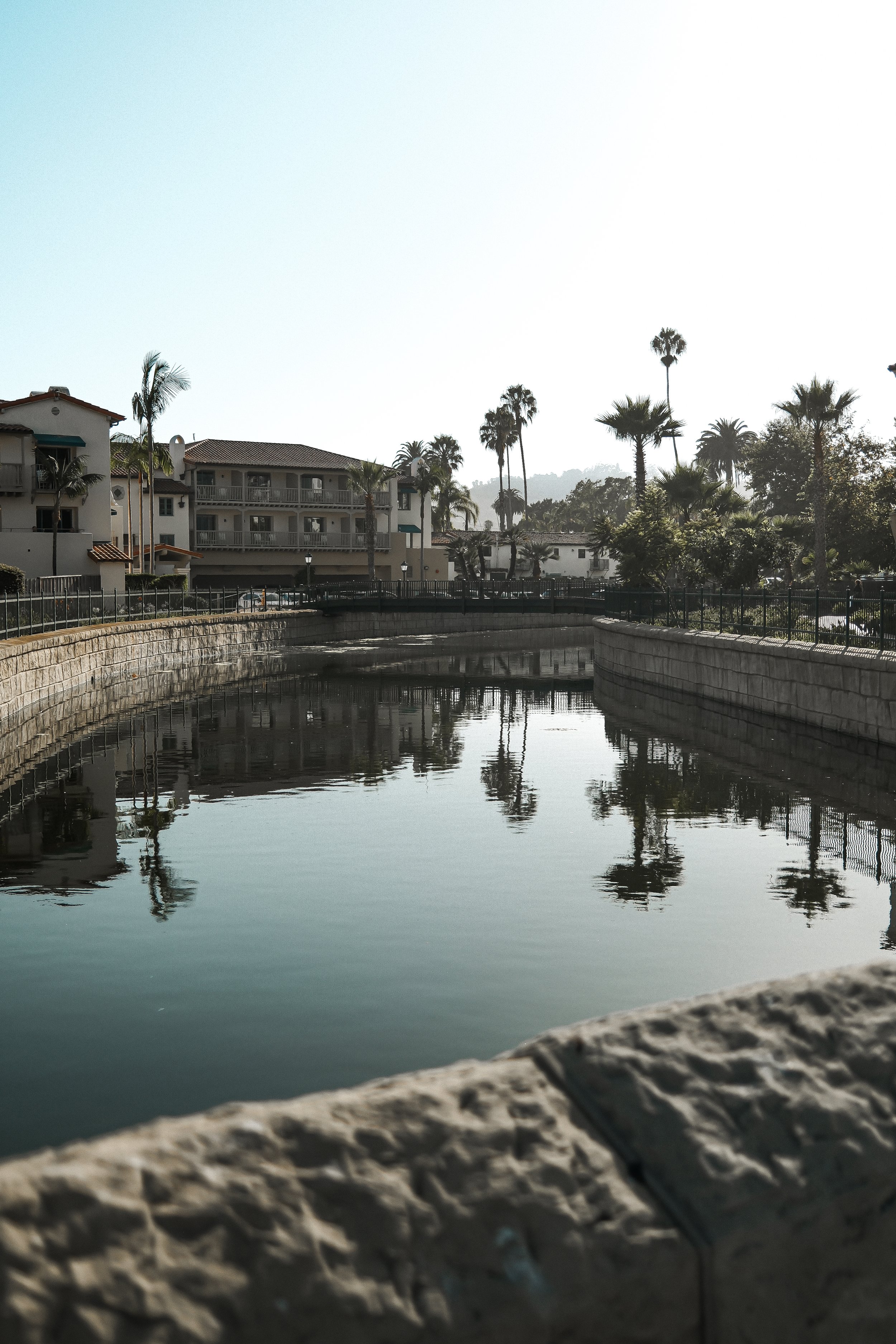 A canal with calm water reflecting residential buildings and palm trees, with a stone wall and a sidewalk in the foreground.