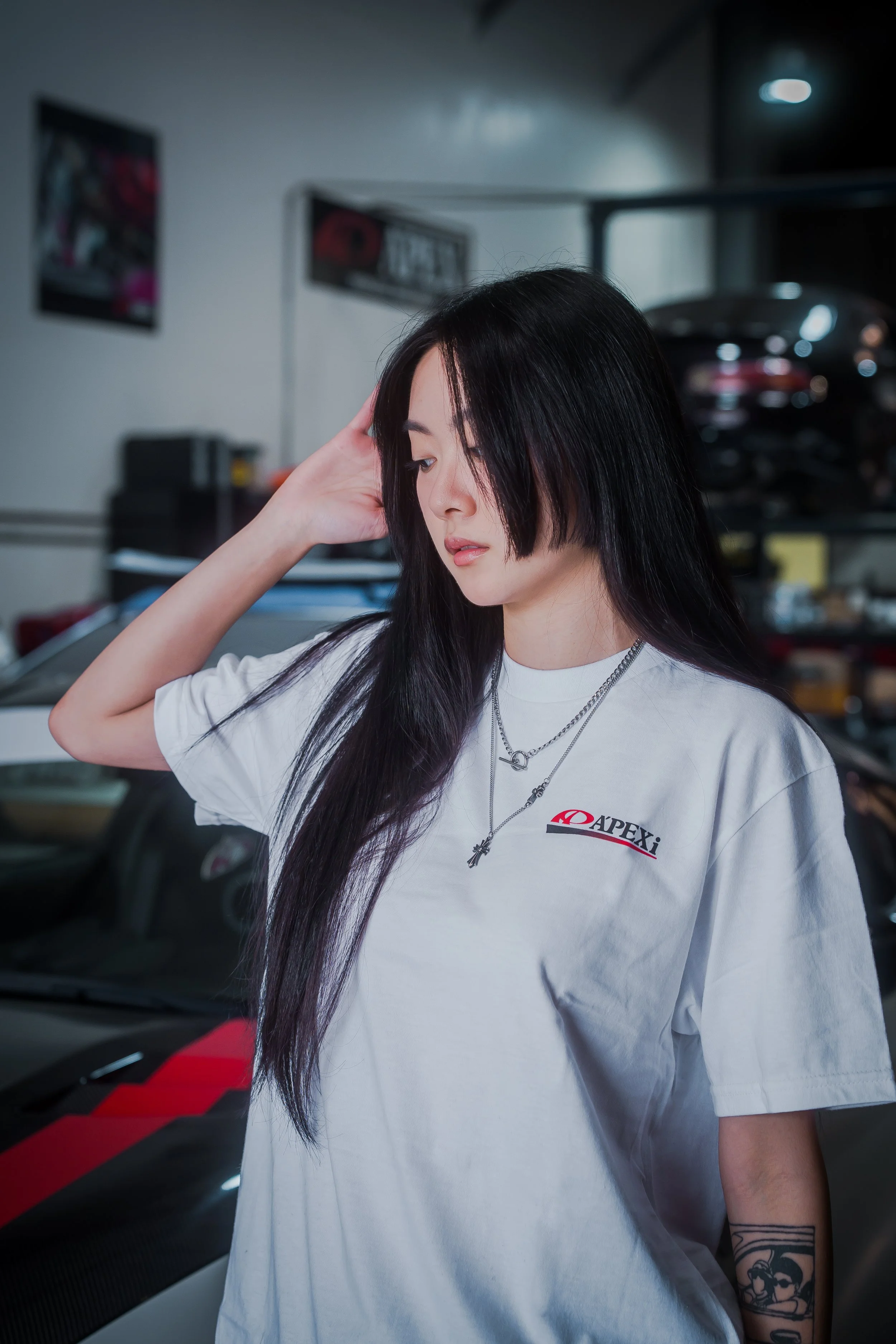 A young woman with long black hair and tattoos on her arm standing in a garage, wearing a white T-shirt with a logo, and looking downward with her hand on her head.