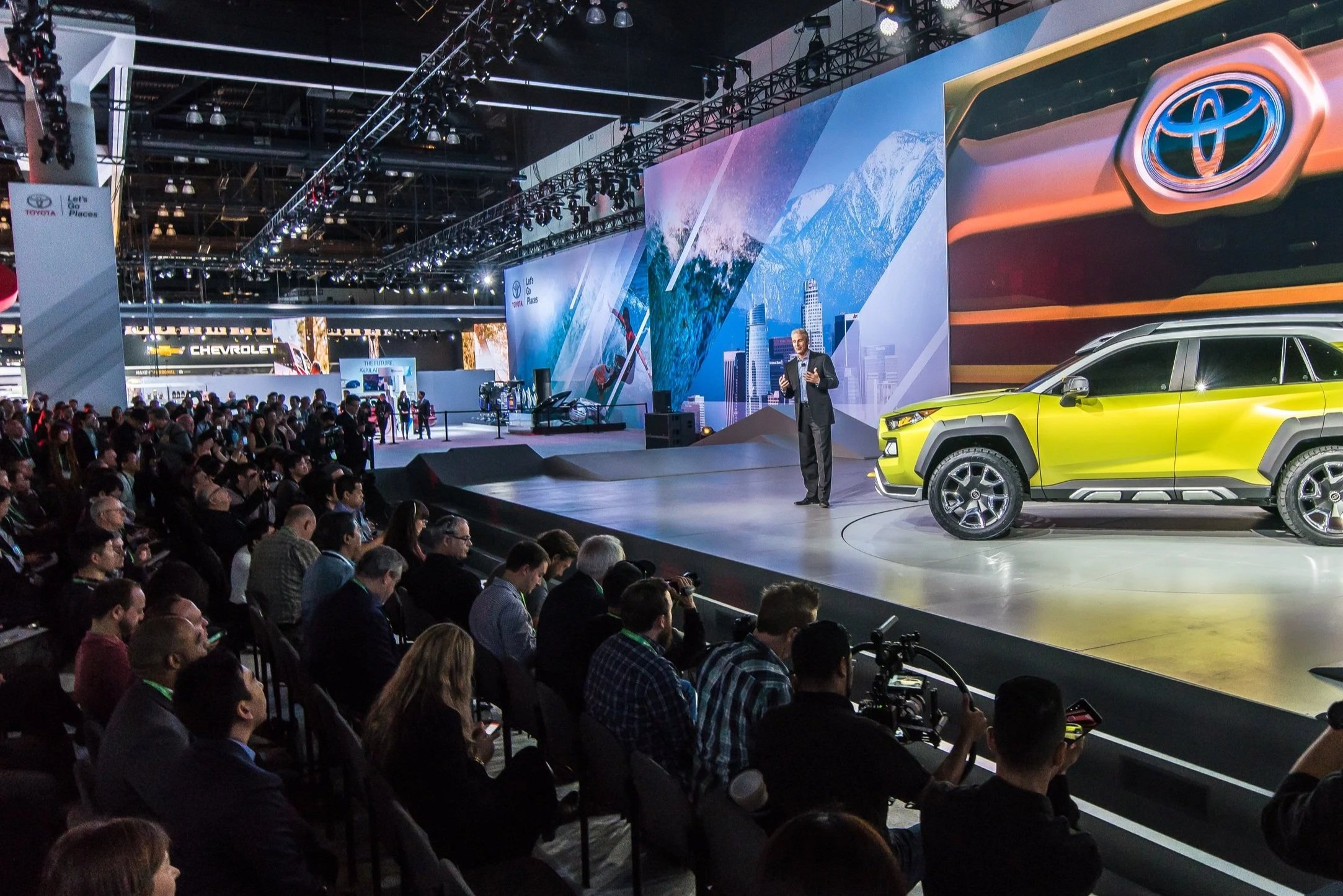 A large audience watching a presenter on stage at a car exhibition. The stage features a yellow SUV and a large backdrop with images of snowy mountains and city buildings. There are displays of car brands, including Toyota and Chevrolet.