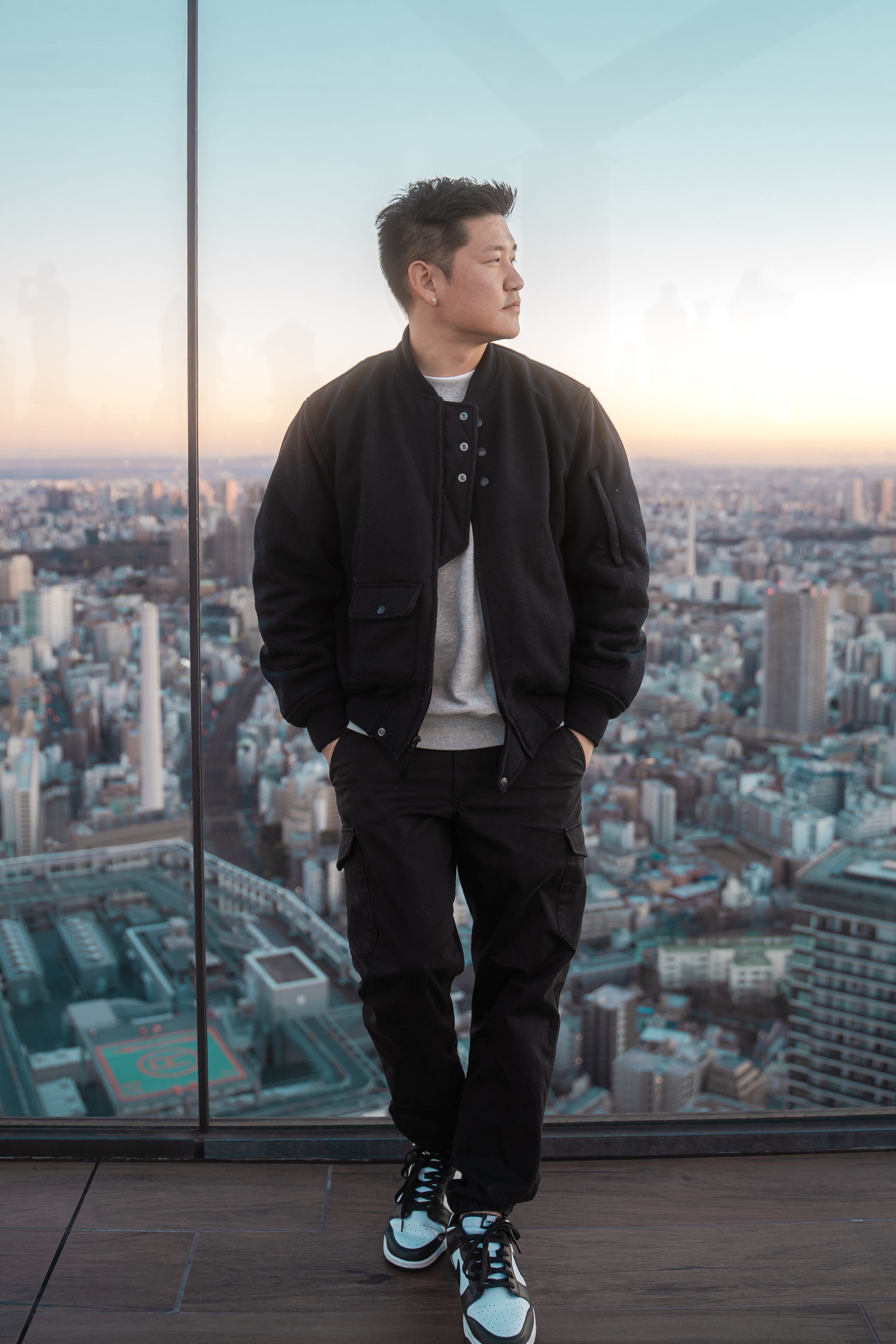 A young man with short dark hair, wearing a black jacket, gray shirt, black pants, and black and white sneakers, stands with hands in pockets on a high rooftop overlooking a cityscape during sunset. The city has many high-rise buildings and a sports court visible below.