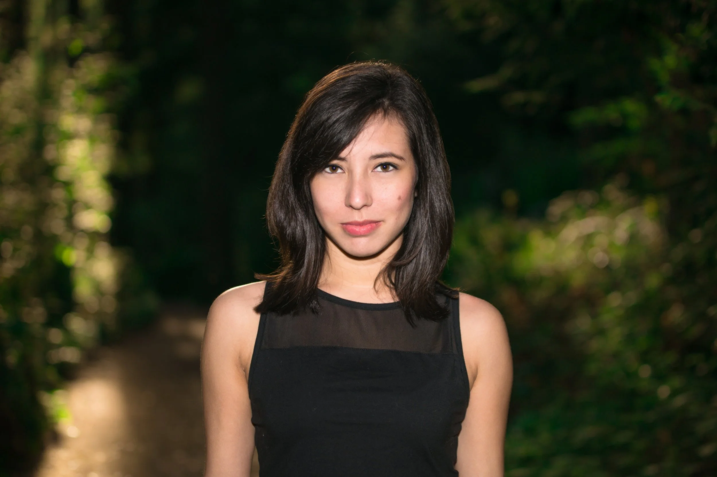 A woman with shoulder-length dark brown hair, wearing a sleeveless black top, standing outdoors in a natural setting with trees and sunlight in the background.