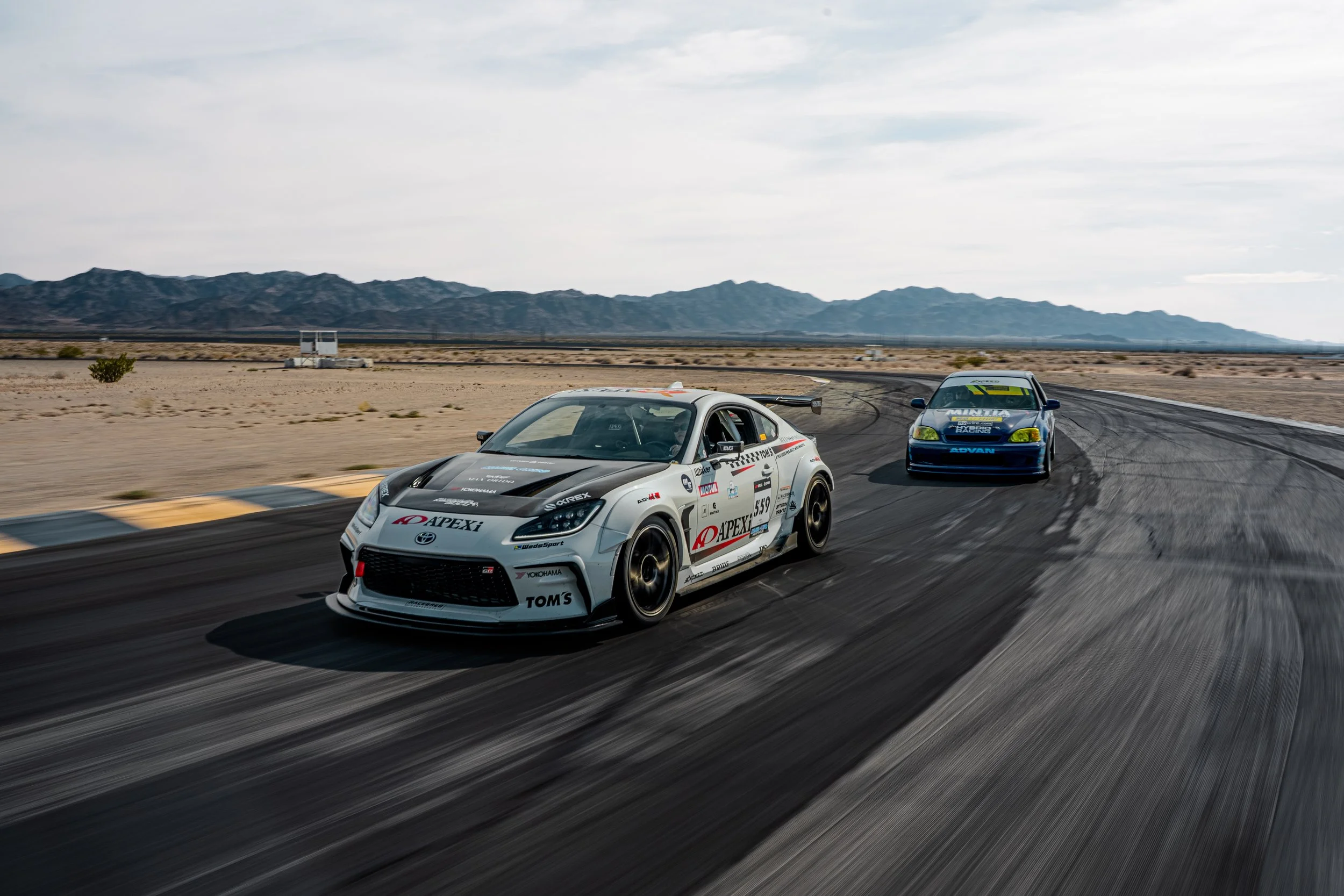 Two cars racing on a desert track with mountains in the background.