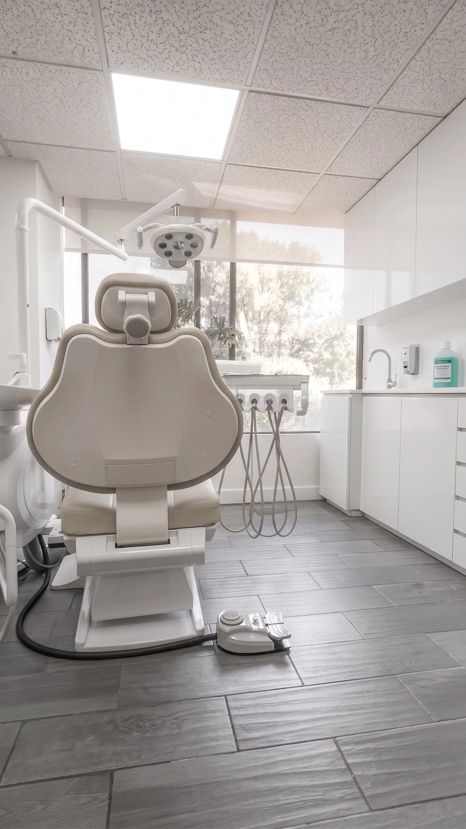 A clean, modern dental clinic with a dental chair, overhead light, and dental tools on a nearby table. Large window with natural light and gray wood-look tiled floor.