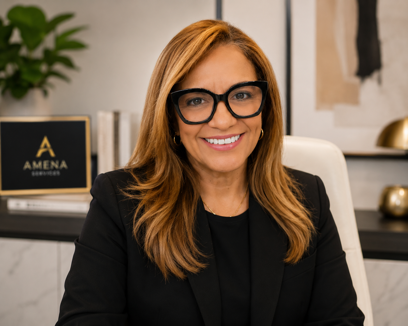 A woman with long red hair and large black glasses, wearing a black blazer, sitting at a desk in an office space, smiling at the camera.