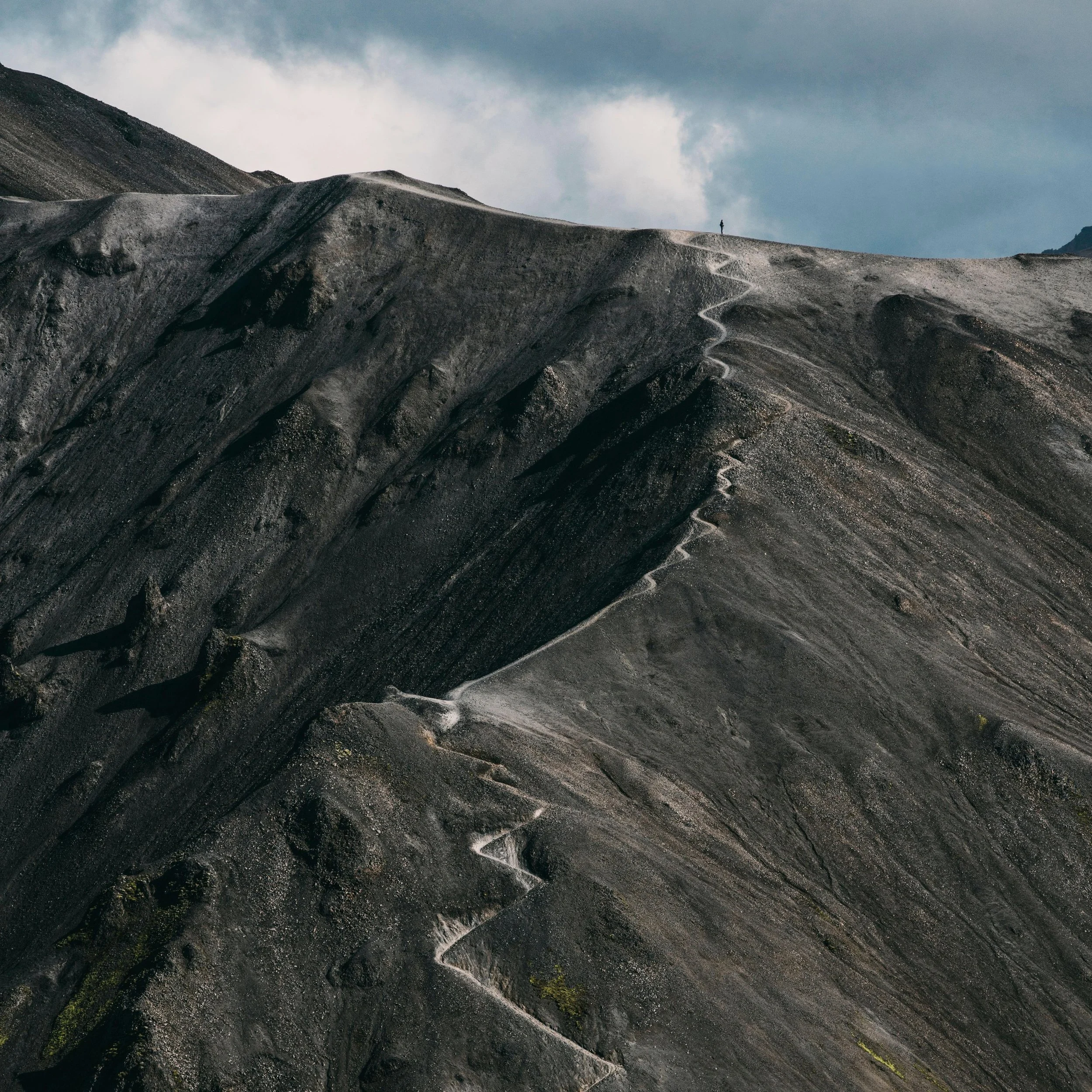 A barren, rocky mountain landscape with a narrow winding trail and a single person walking along the ridge under a cloudy sky.