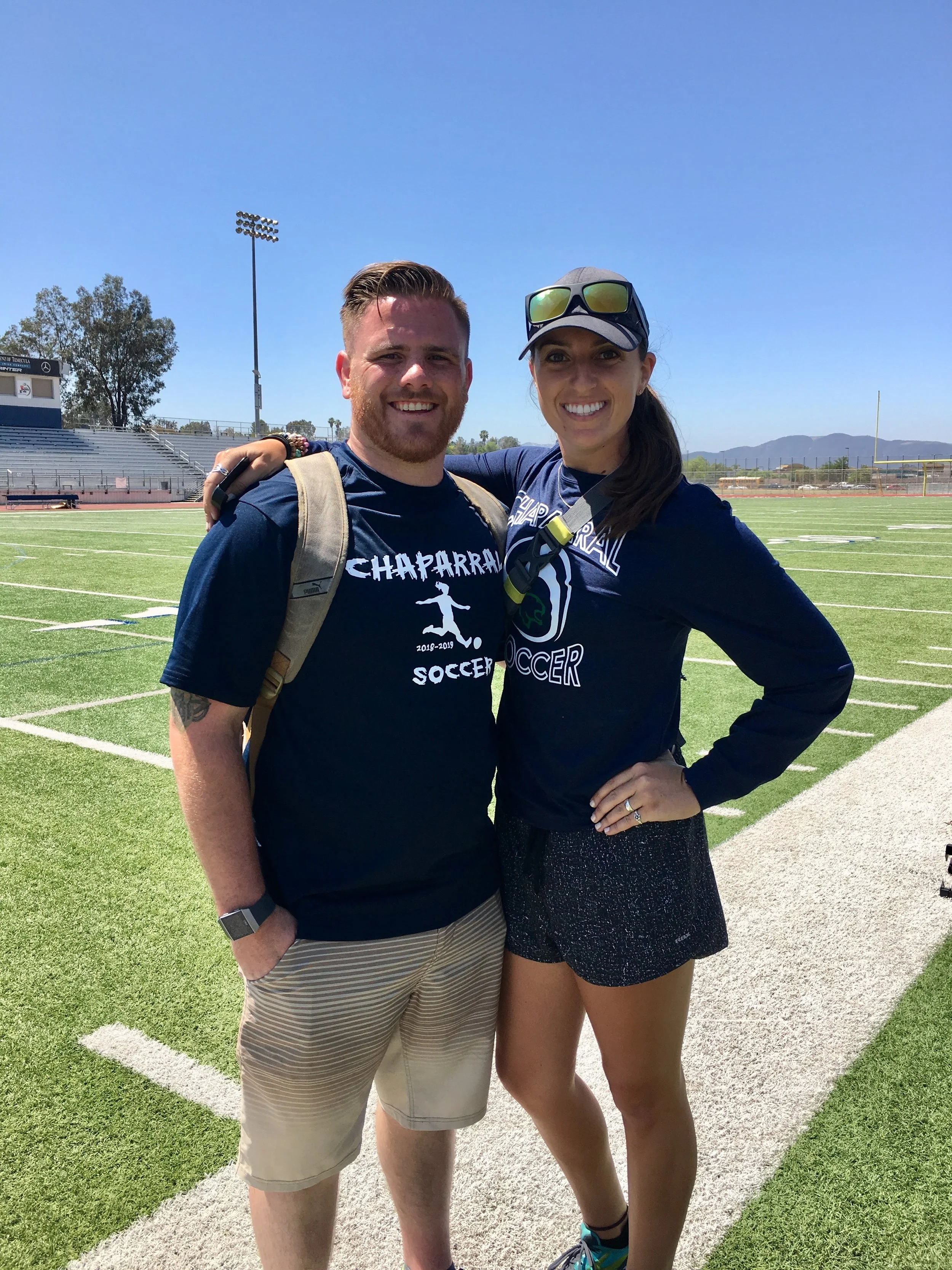Two smiling athletes, a man and a woman, standing on a football field with stadium seats and goalpost in the background under a clear blue sky.