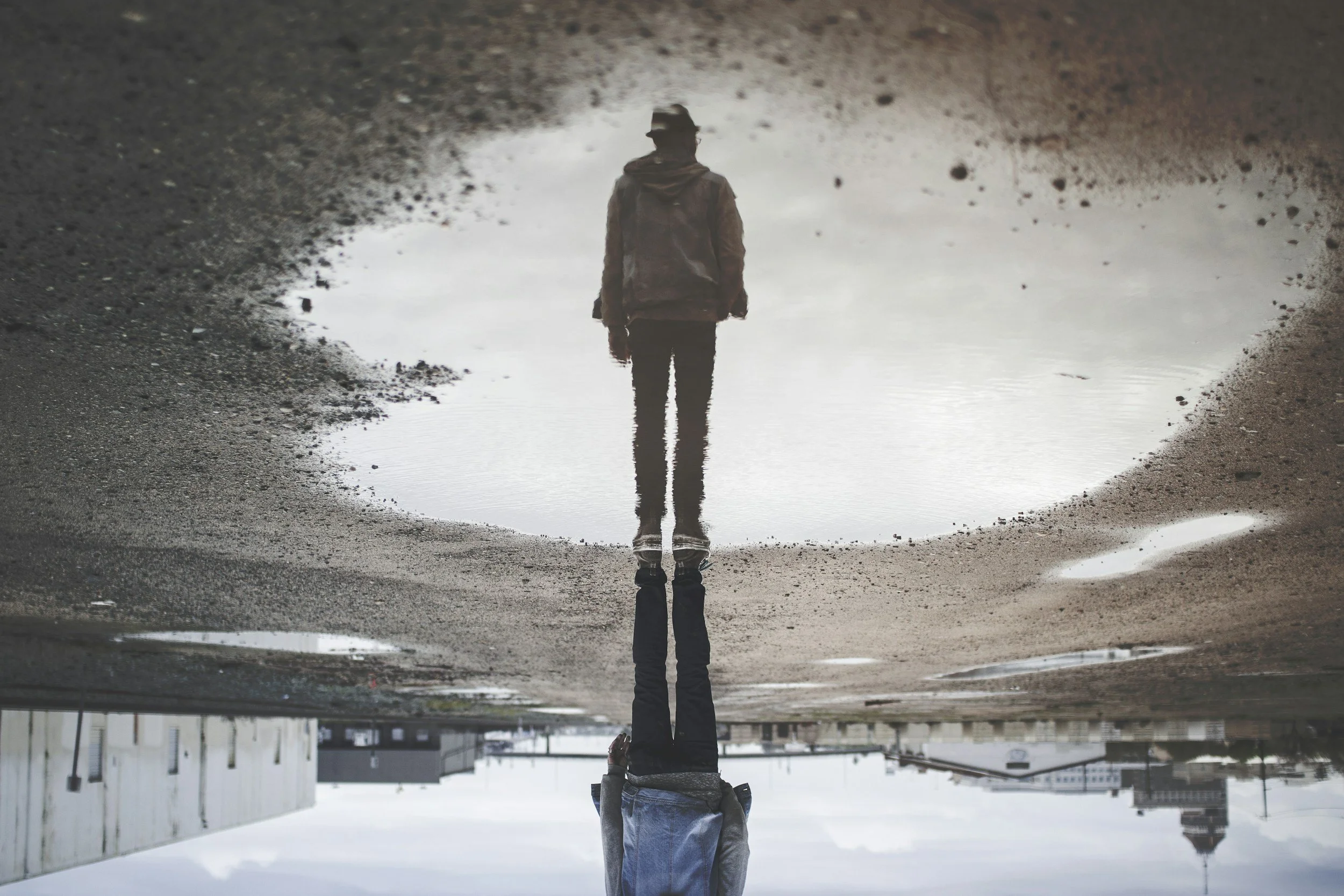 A person standing in a puddle of water on the ground, viewed upside down, with the sky and buildings reflected in the water.