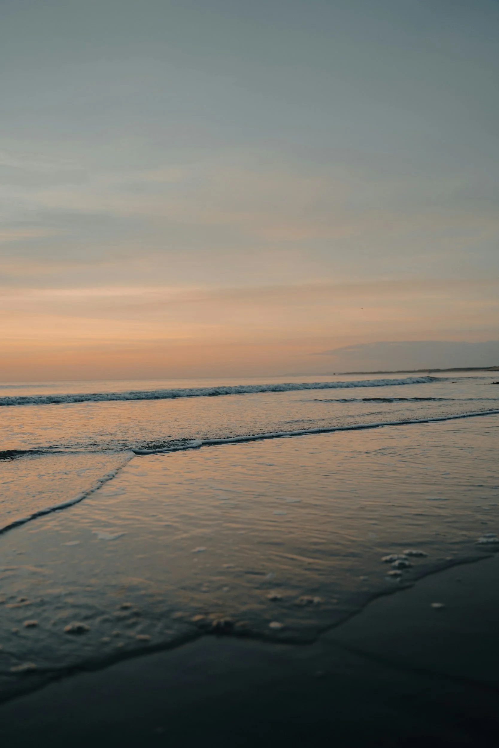 A serene beach scene during sunset with calm waves gently washing onto the shore, sky painted with pastel hues of orange, pink, and blue.