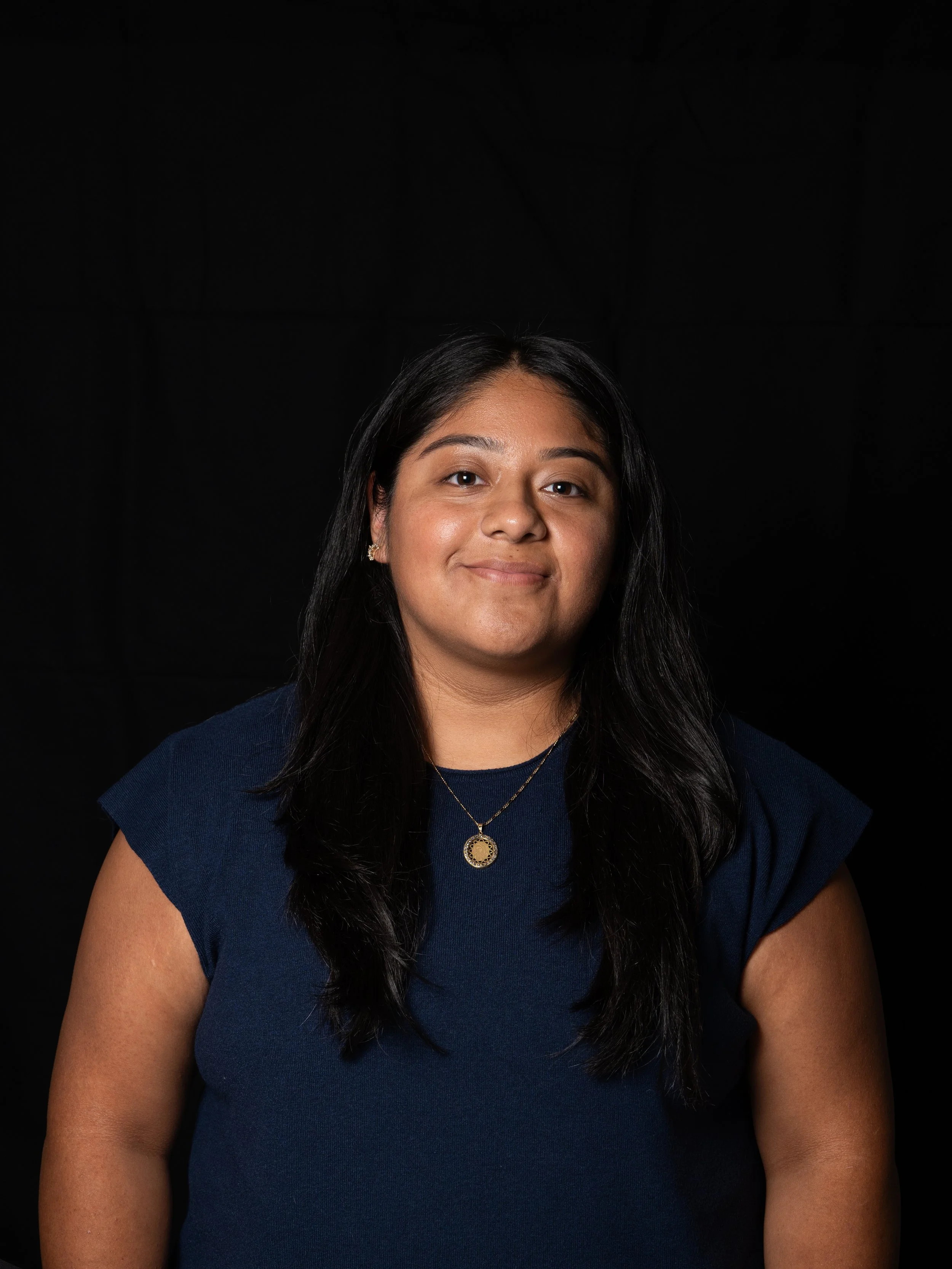 A young woman with long black hair, wearing a navy blue top, gold earrings, and a gold pendant necklace, smiling against a black background.