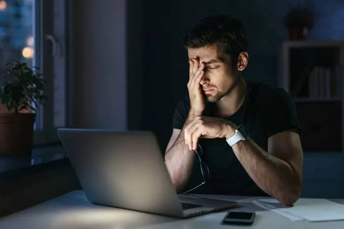 A man sitting at a desk in a dark room, holding his forehead in frustration or fatigue, with a laptop open in front of him, and a smartphone nearby.