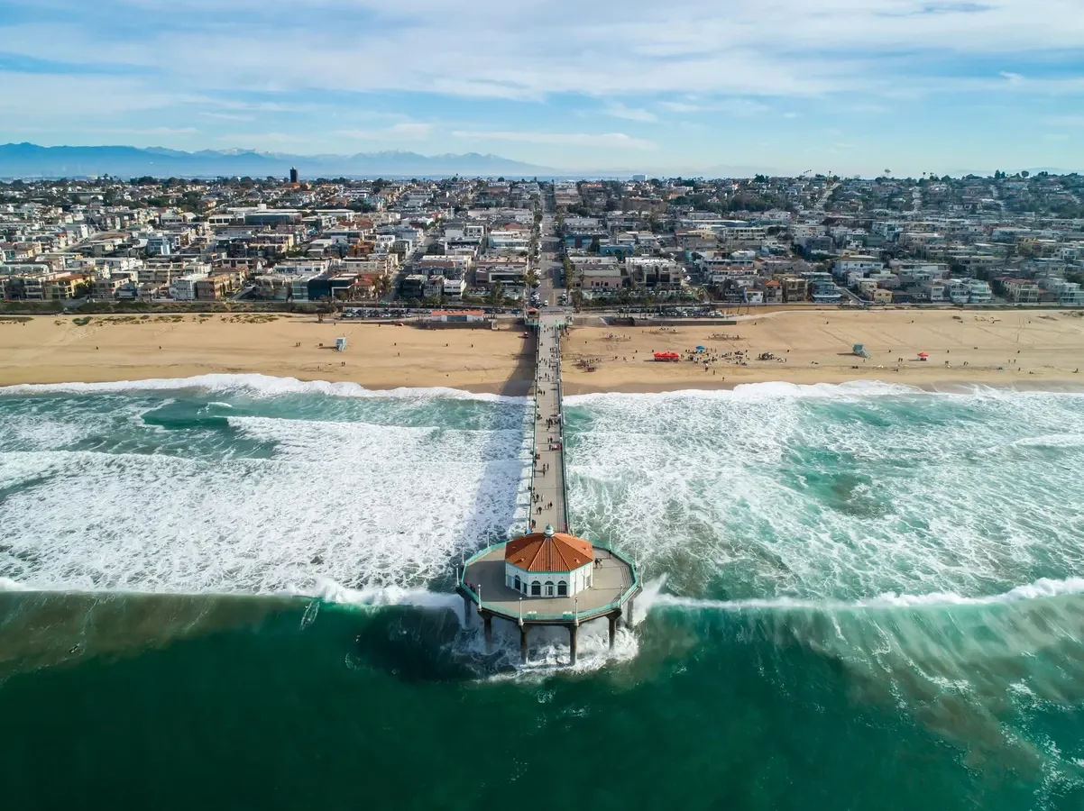 Aerial view of a pier extending into the ocean in front of a city and beach with waves crashing on the shore.