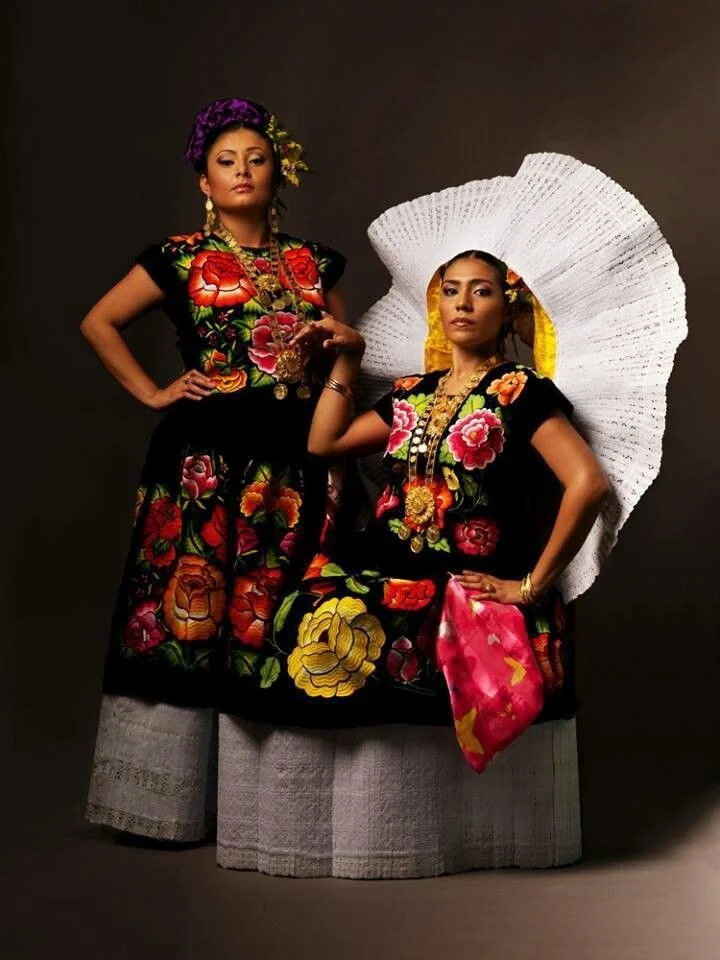 Two women wearing traditional Mexican dresses with colorful floral embroidery, one with a large white sombrero-style accessory and the other with a purple flower headpiece, both adorned with jewelry and standing against a dark background.