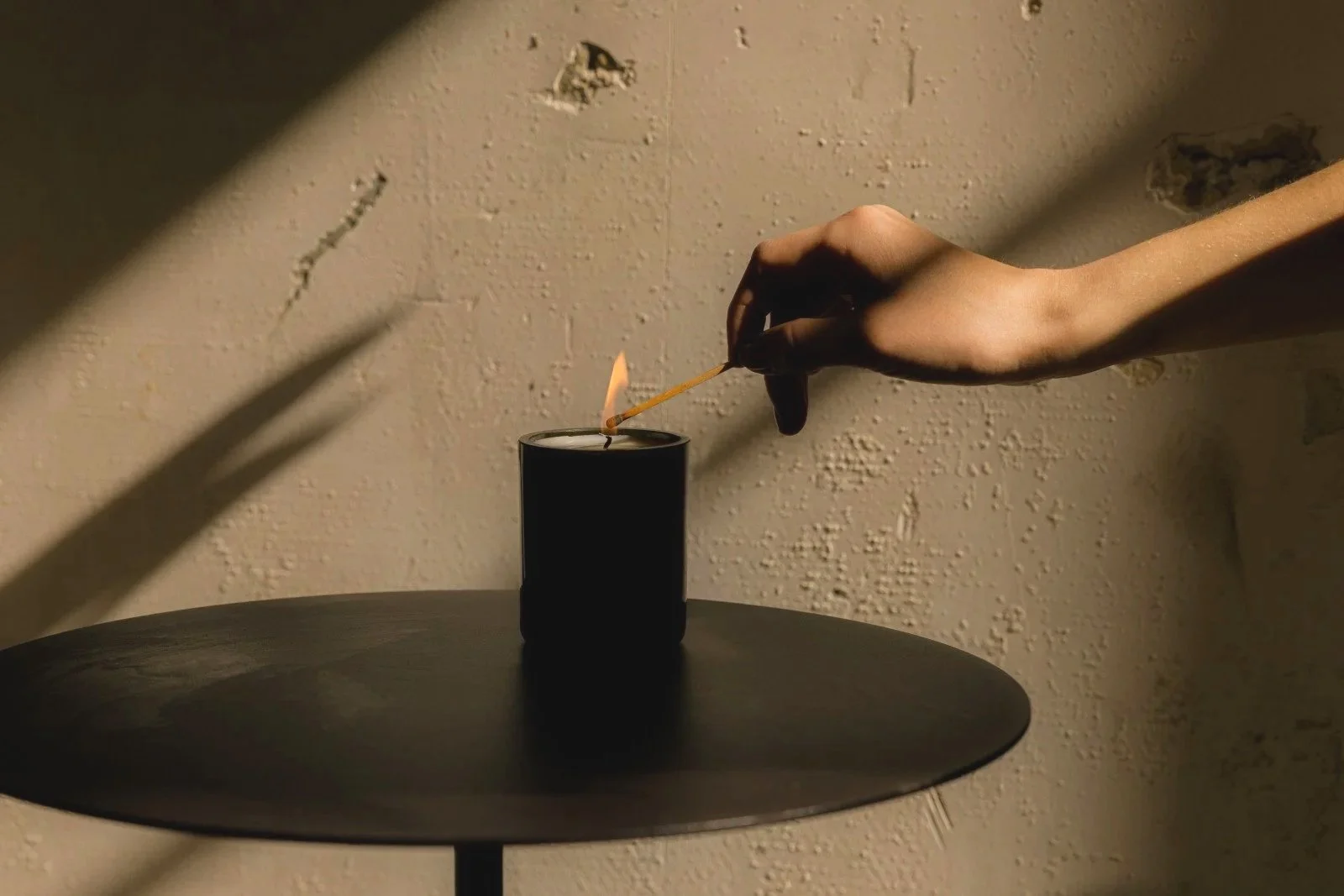 A close-up of a person's hand lighting a black candle with a matchstick in a dimly-lit room. The background is a beige textured wall.