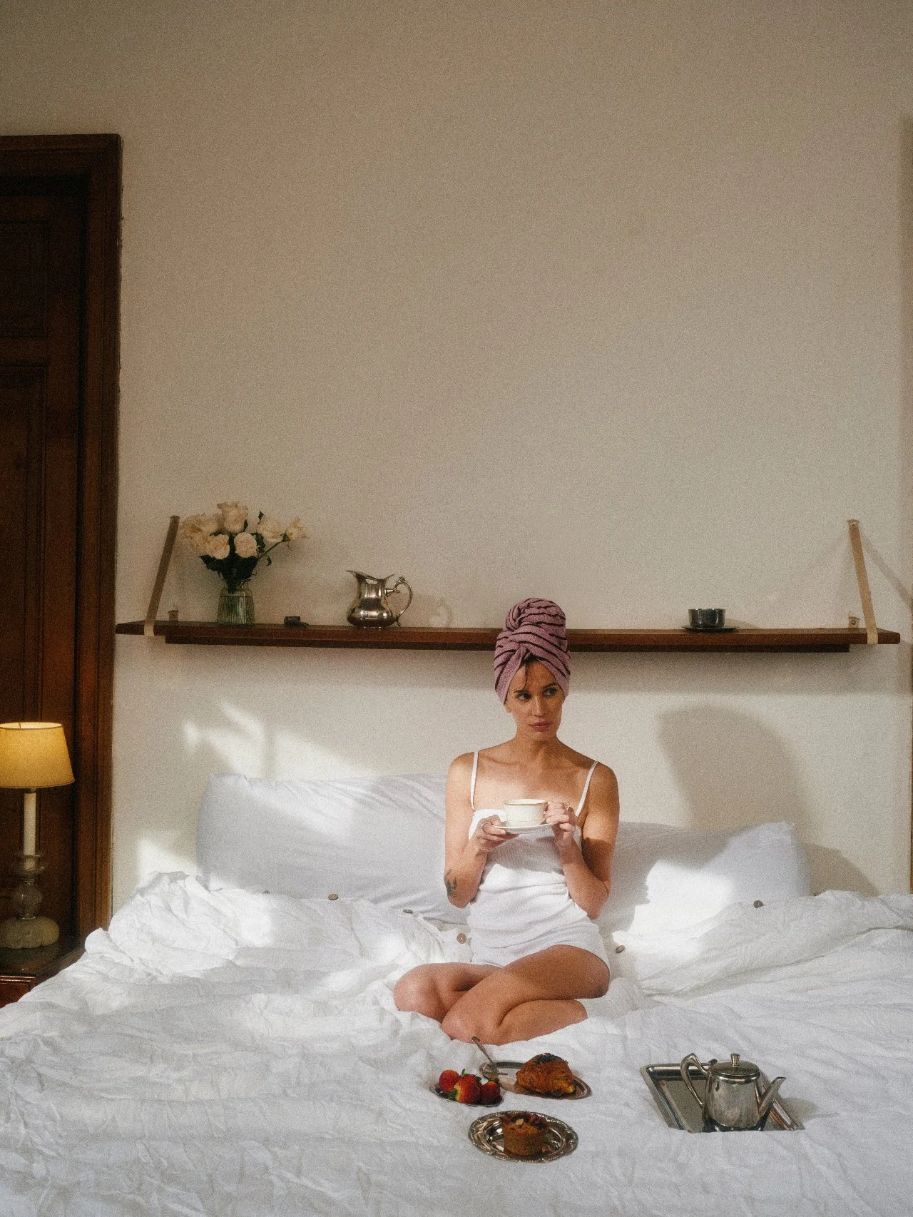 A woman sitting on a bed with a towel wrapped around her head, holding a teacup, with breakfast items on the bed and a floating shelf with flowers and decorative items behind her.
