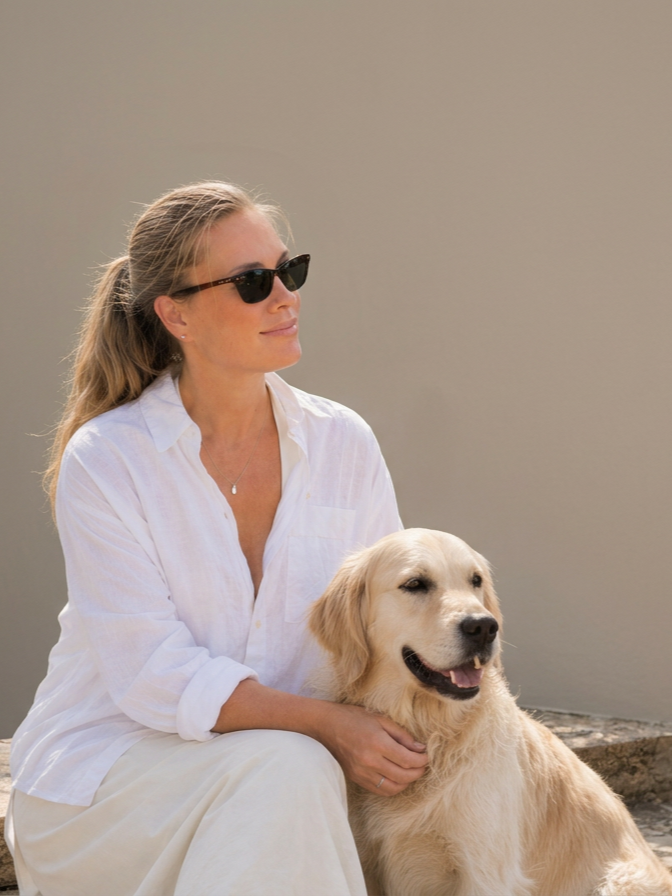 A woman wearing sunglasses and a white shirt sits outdoors next to a golden retriever dog.