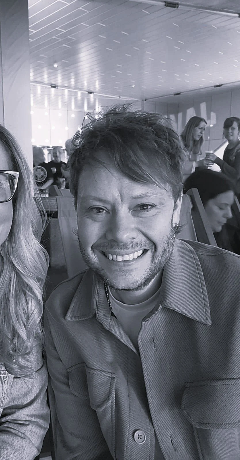 Black-and-white selfie of a smiling man in a collared coat and people mingling in a bright indoor event space.
