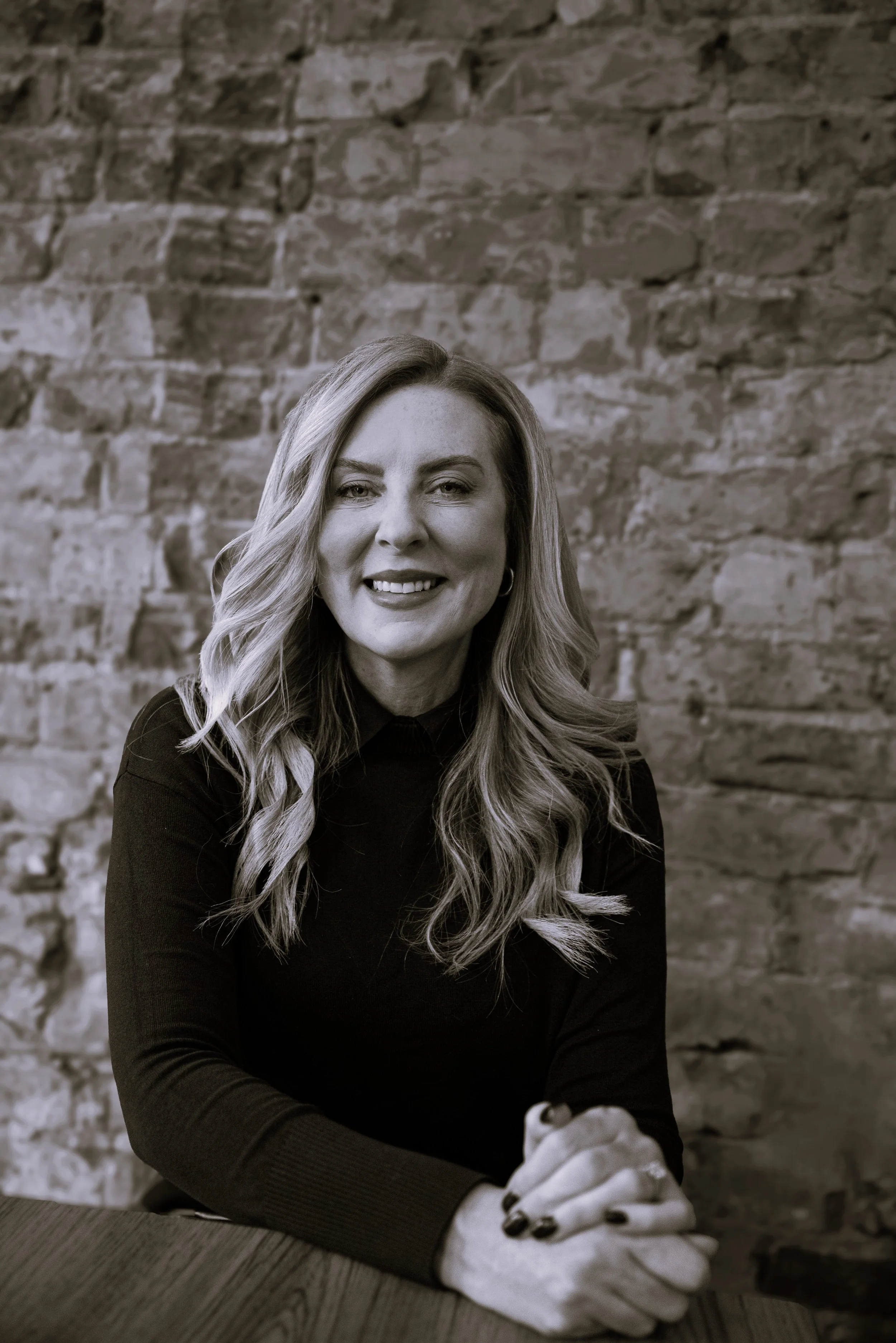 Black-and-white portrait of a smiling woman with long wavy hair, seated at a wooden table with hands folded against a brick wall; warm, confident mood.