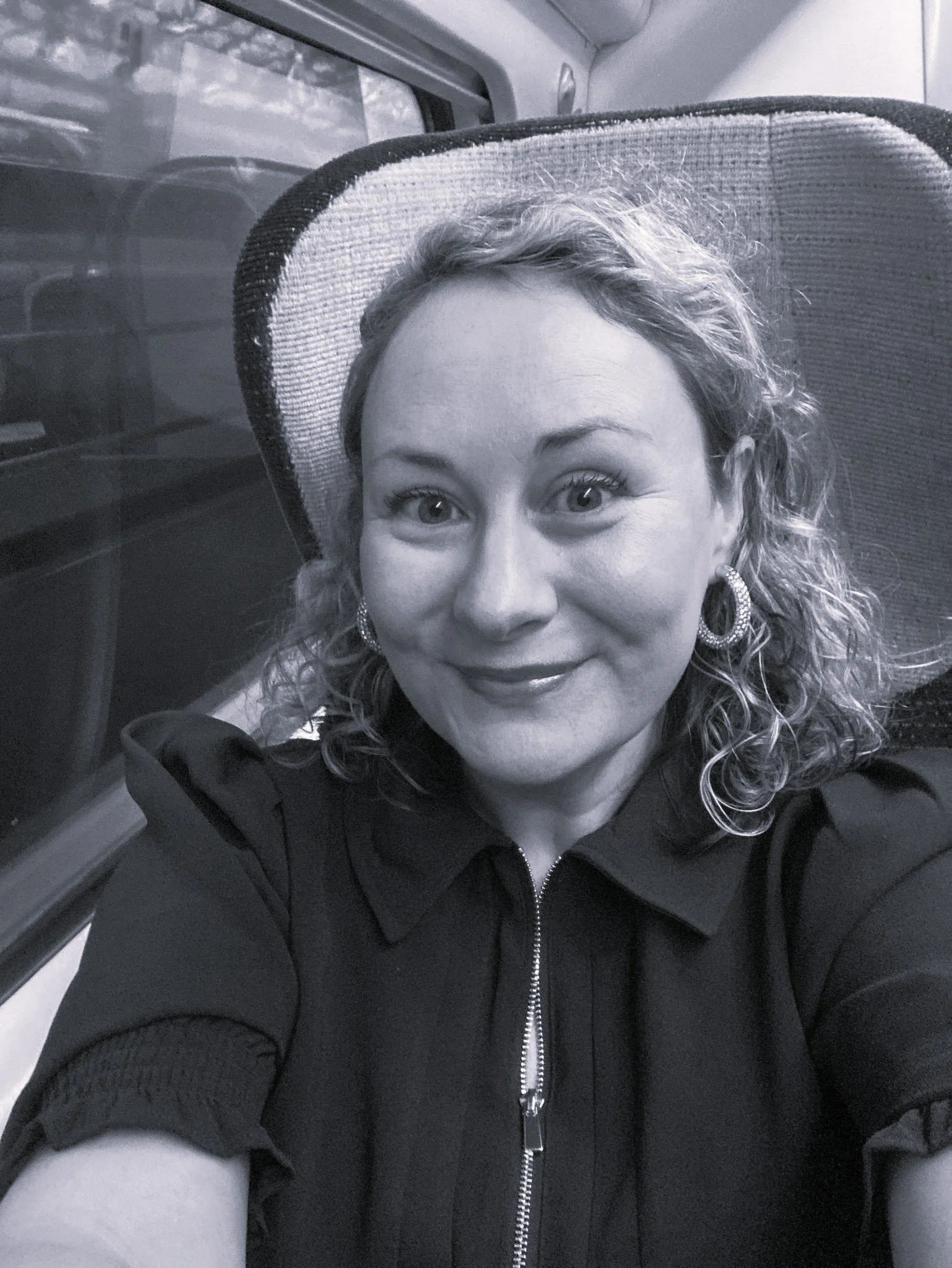 Black-and-white selfie of a smiling woman with curly hair and hoop earrings seated by a train window; the candid portrait feels warm and relaxed.