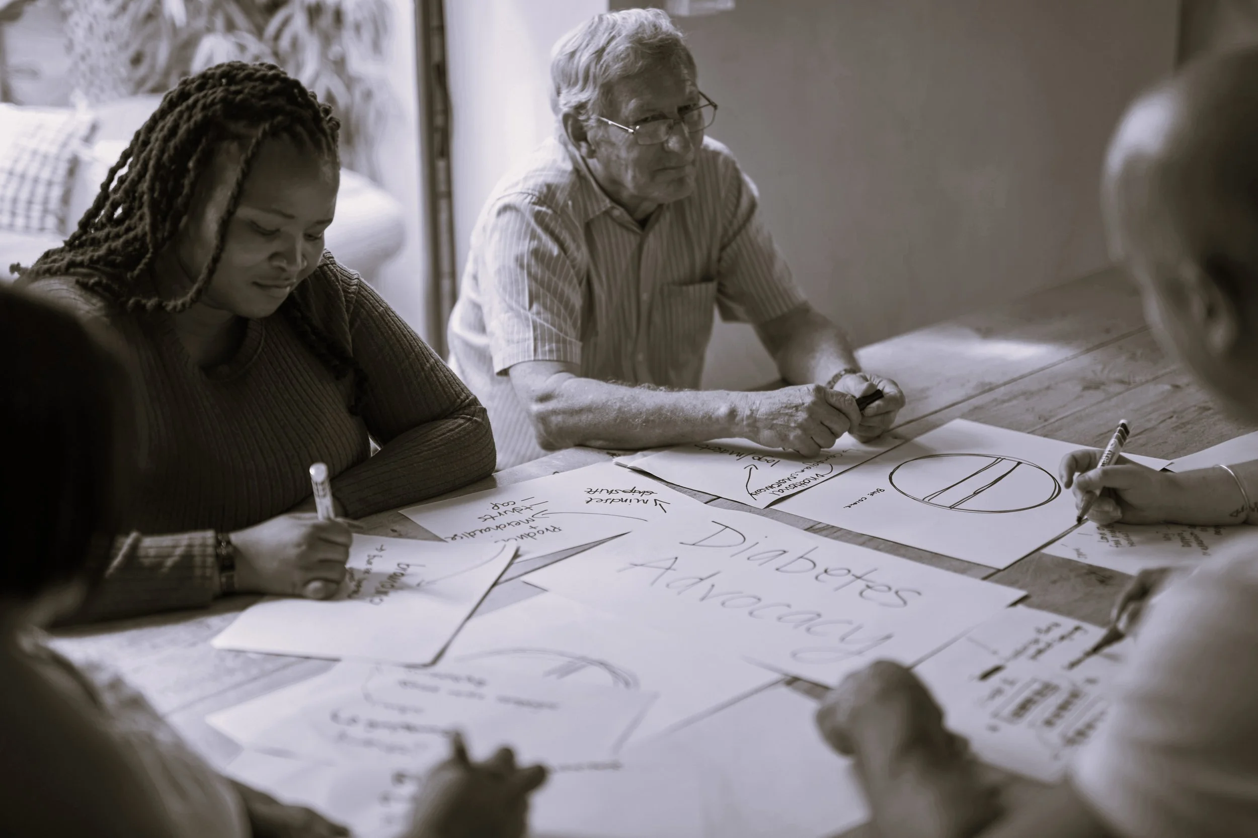 A diverse group of adults collaboratively writes and sketches ideas on large sheets of paper around a table, conveying focused, thoughtful teamwork.
