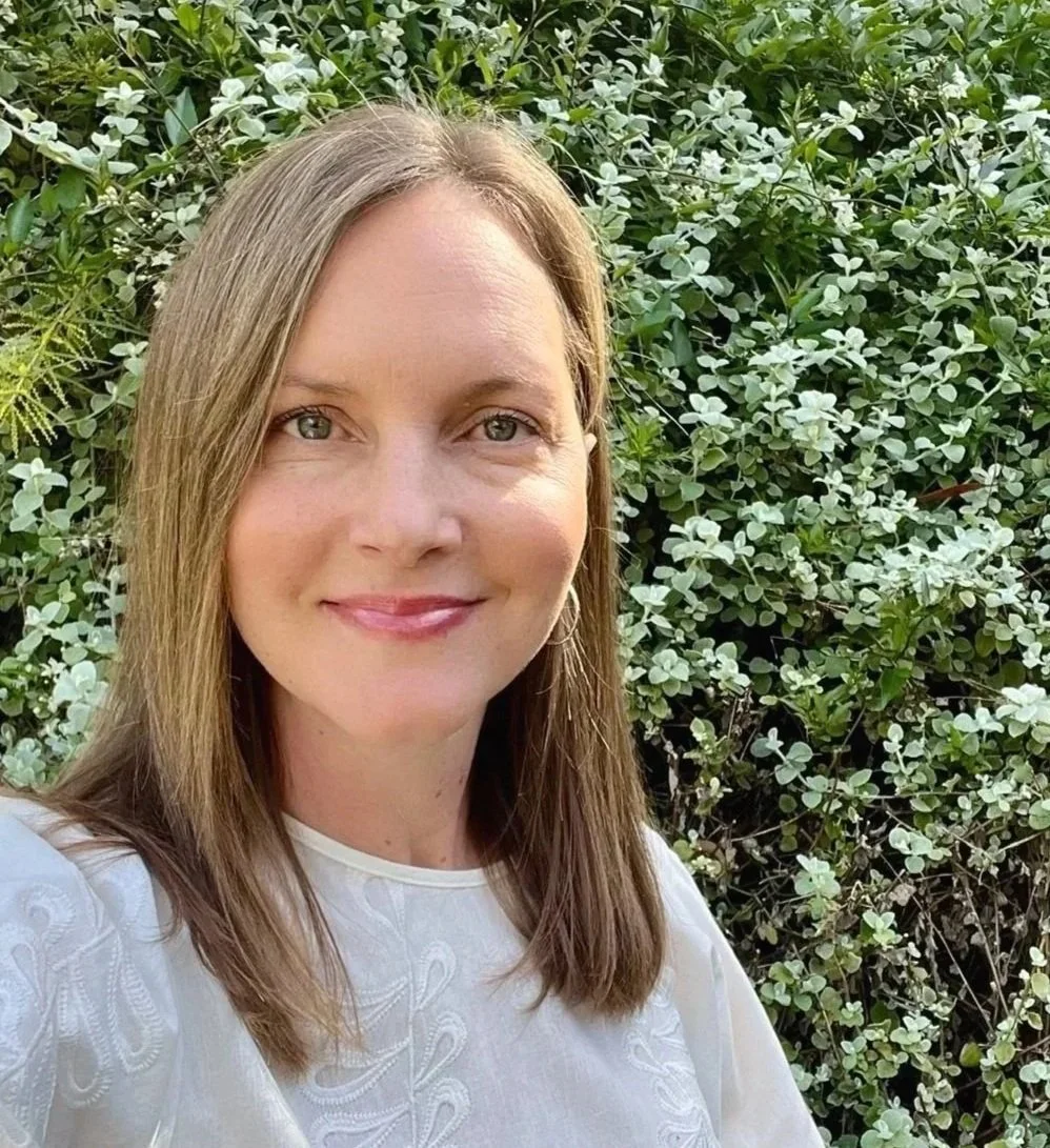 A woman with shoulder-length light brown hair and light skin smiling in front of green leafy bushes.