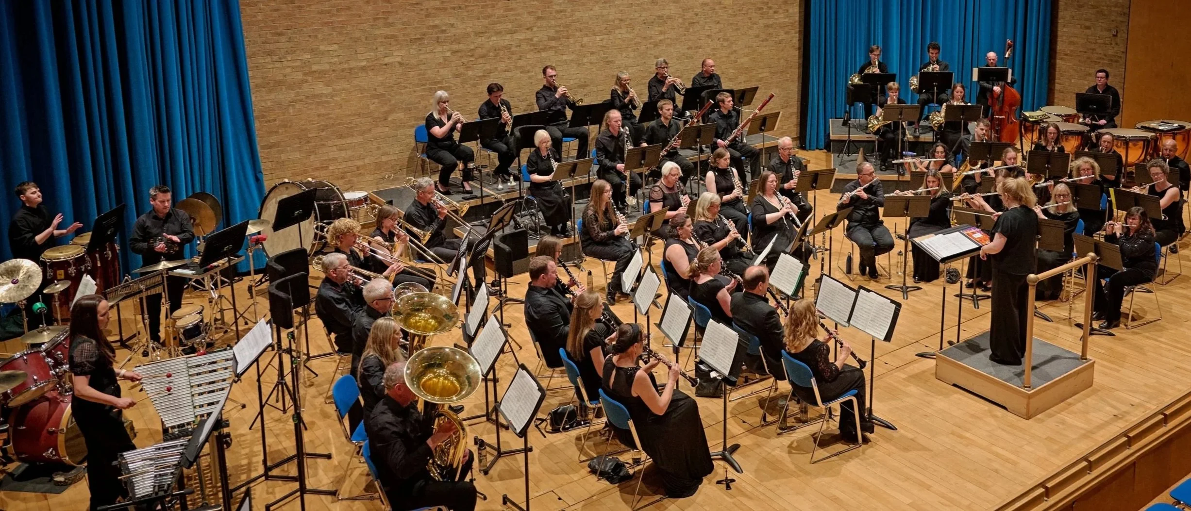 Orchestra performing on stage in concert hall with conductor leading, blue curtains and brick wall background, various musicians playing instruments including clarinets, saxophones, drums, and percussion.