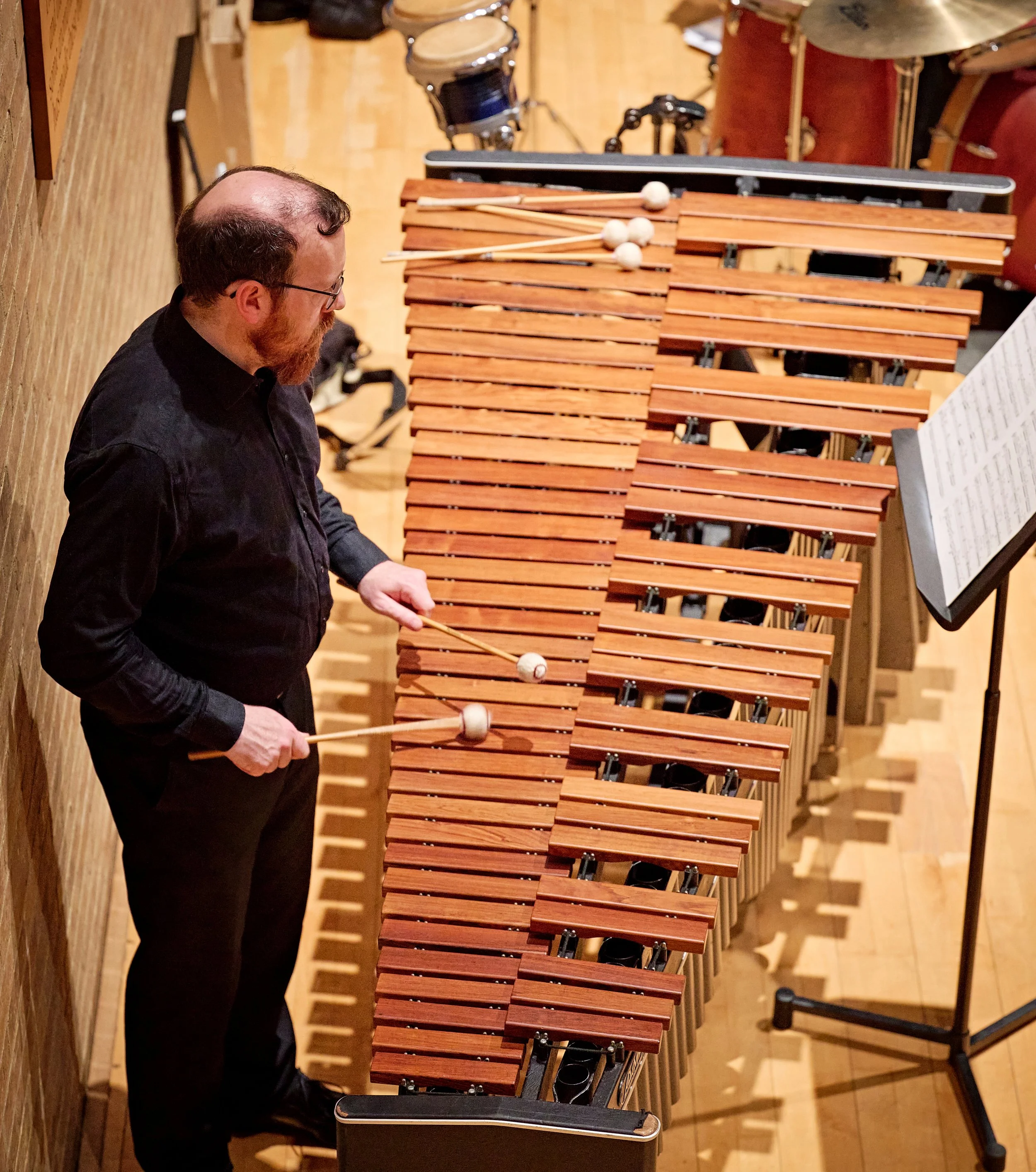 A man with glasses and a beard playing a marimba, positioned against a wooden wall in a concert hall. The marimba has wooden bars with mallets resting on top, and sheet music is on a stand nearby.