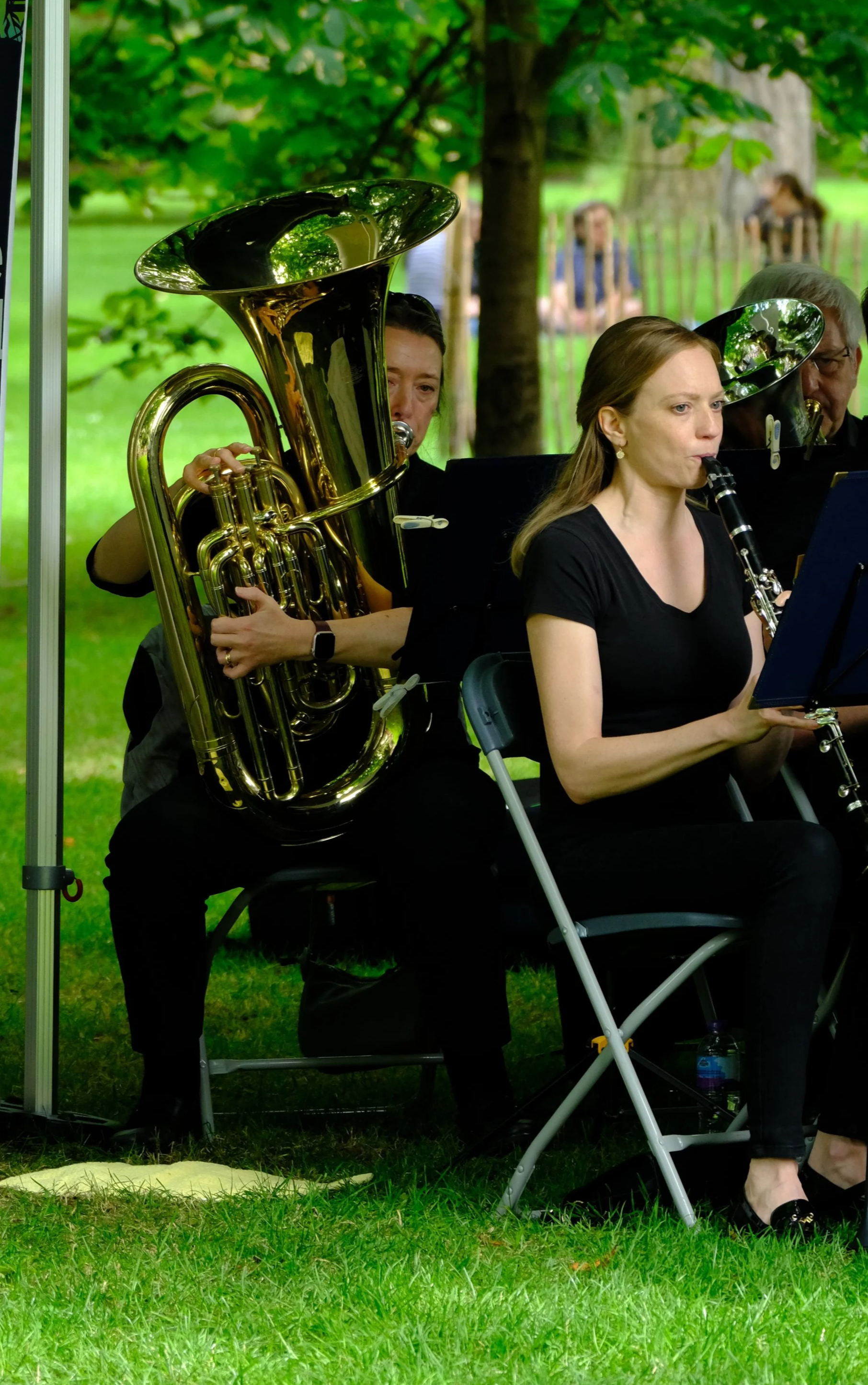 Musicians playing wind instruments outdoors in a park with green trees and grass, seated on black chairs under a canopy.
