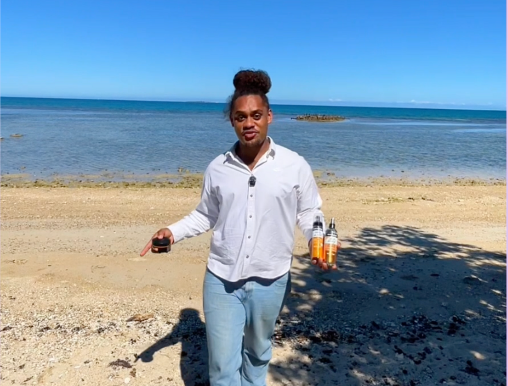 Une femme debout sur la plage, tenant deux bouteilles et un petit pot, avec la mer en arrière-plan et un ciel bleu.