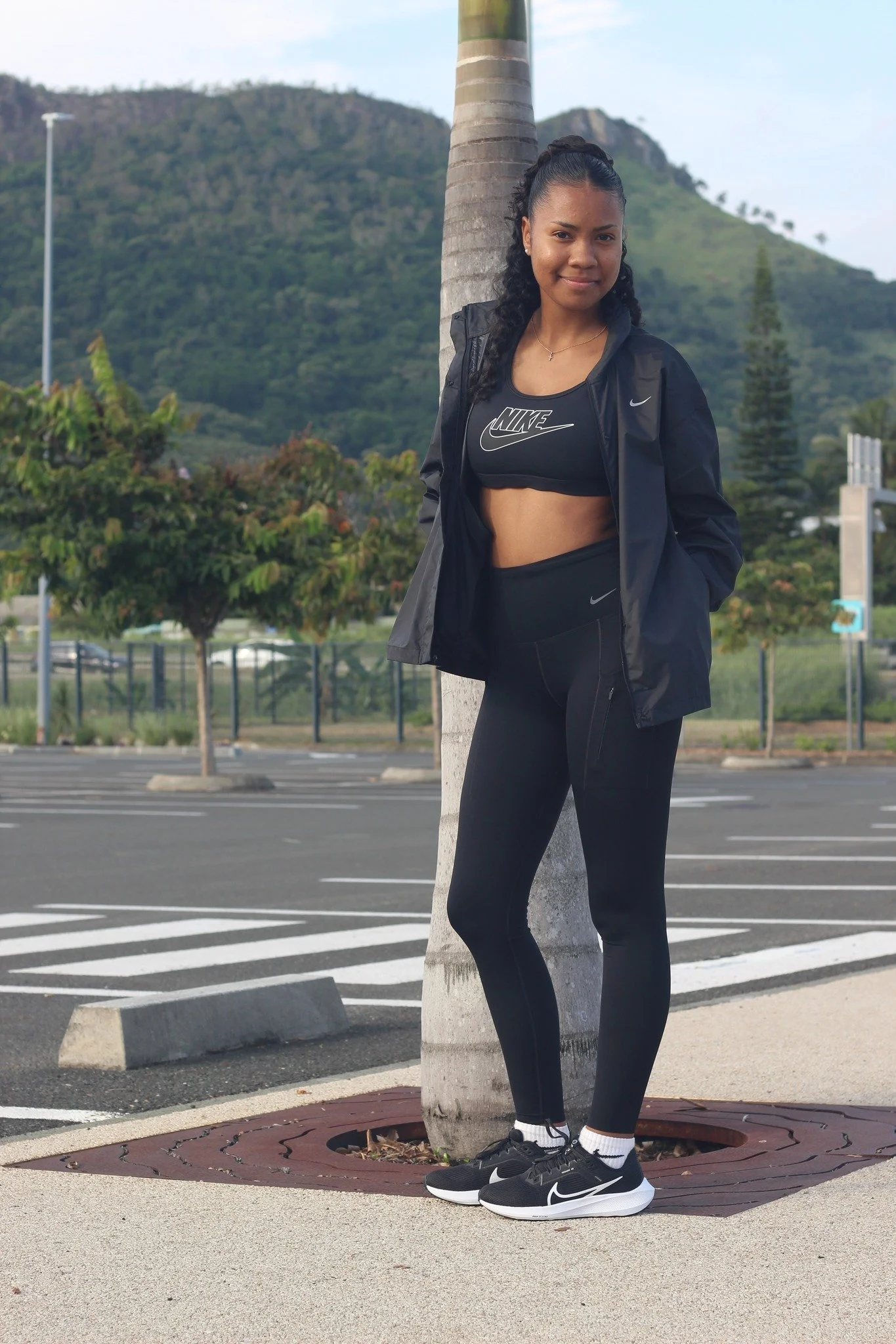 Une jeune femme habillée en vêtements de sport, portant des chaussures de course Nike, se tient contre un arbre dans un parking avec des montagnes en arrière-plan, le jour