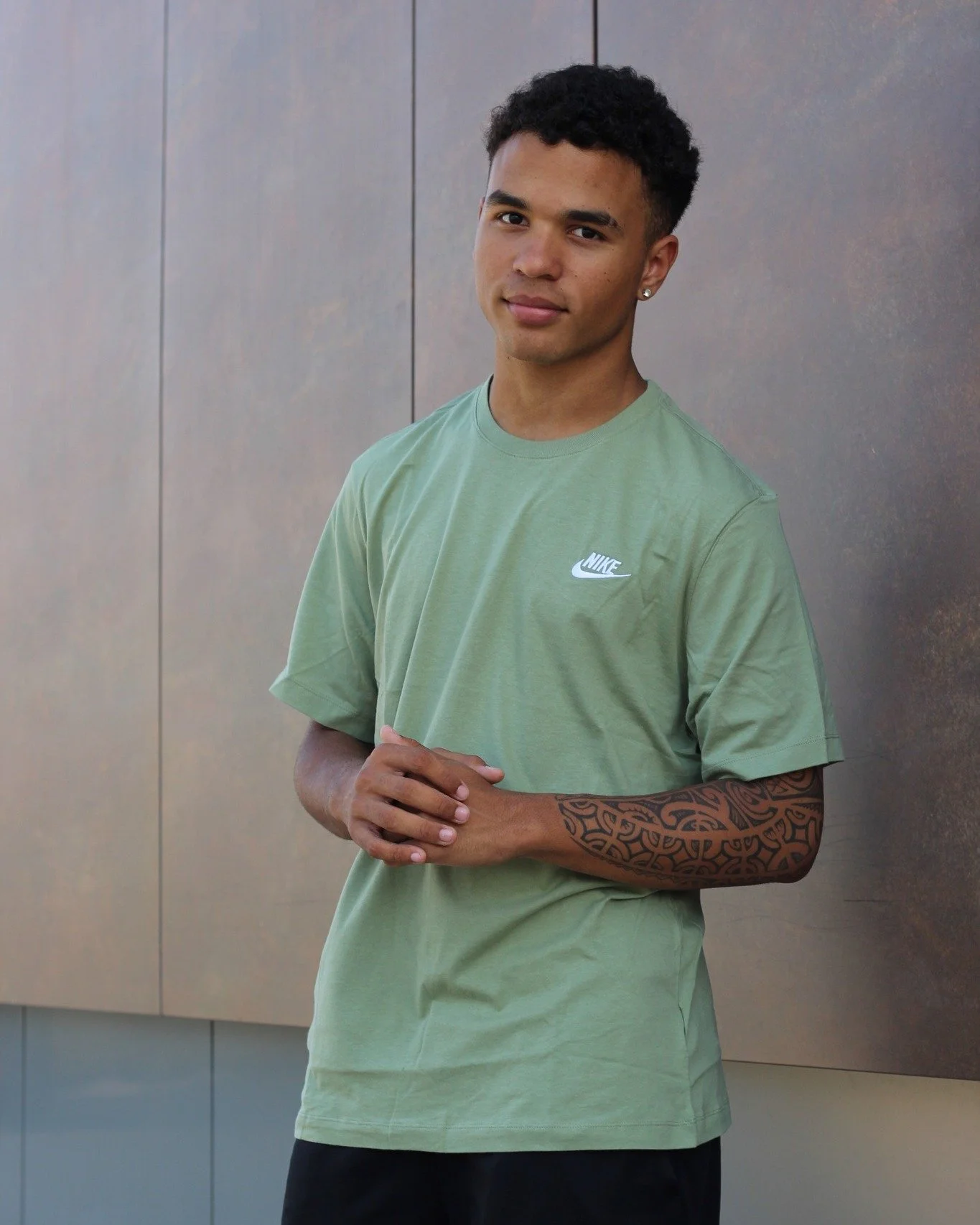 Un jeune homme aux cheveux courts et bouclés porte un t-shirt vert Nike et des boucles d'oreilles argentées, debout contre un mur en métal.
