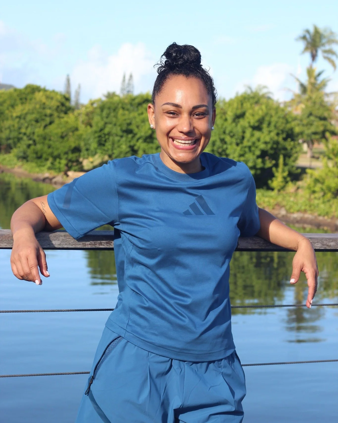 Une femme souriante en tenue de sport bleue, se tenant au bord d'un lac avec des arbres en arrière-plan, sous un ciel clair.
