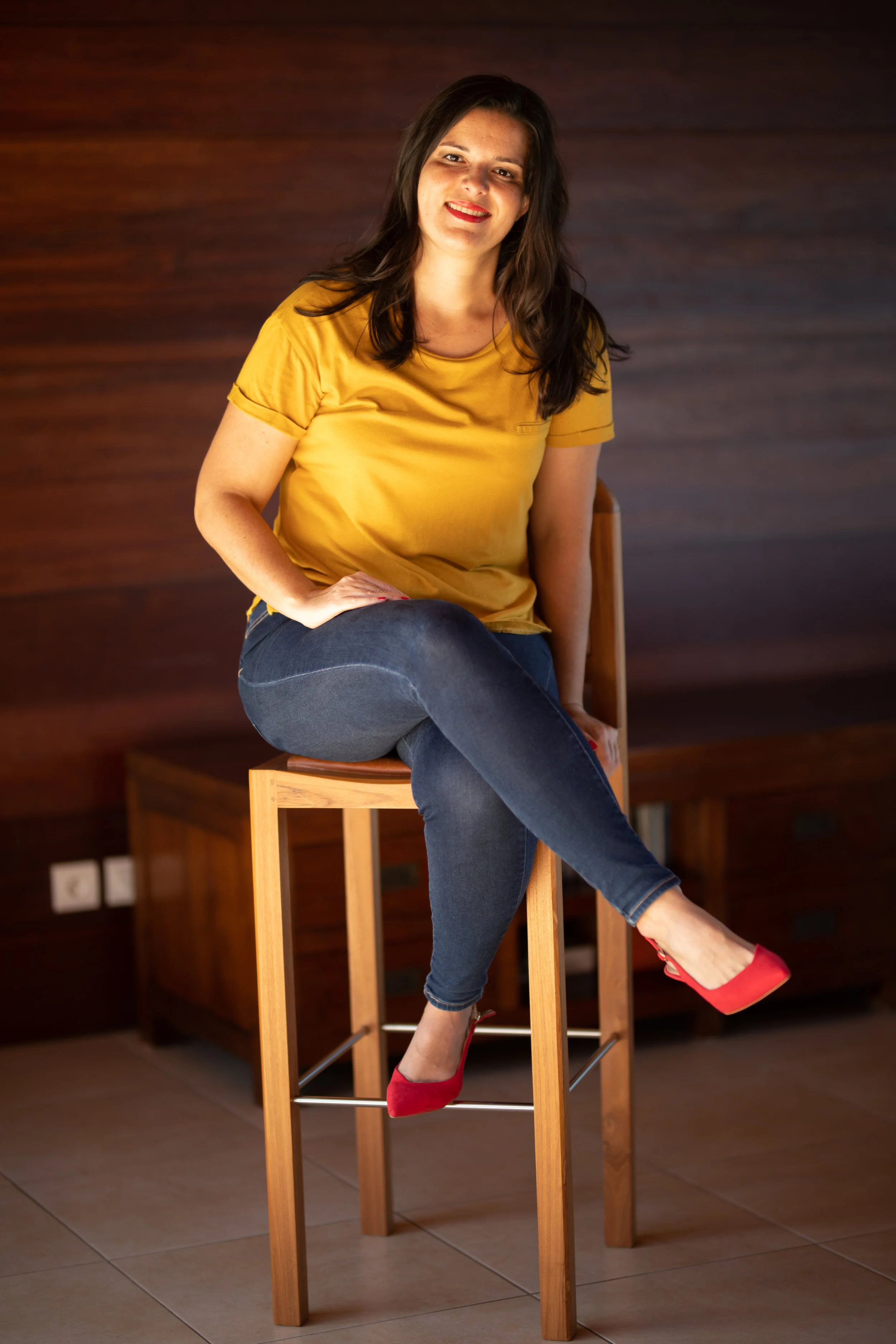 Femme assise sur une chaise haute en bois, portant un t-shirt jaune, un jean foncé, et des chaussures rouges, dans une pièce avec un mur en bois foncé en arrière-plan.