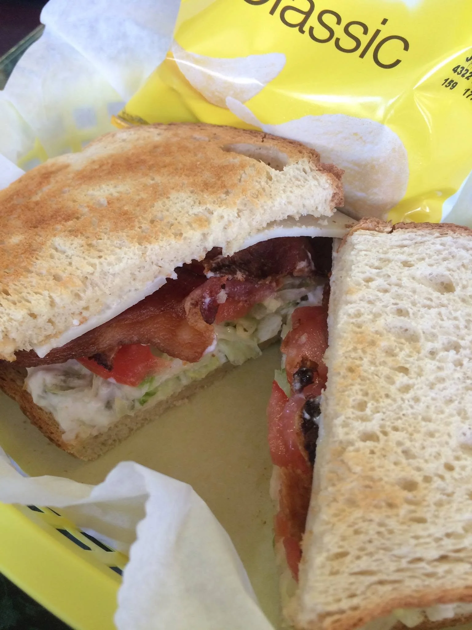 Close-up of a sandwich with bacon, tomato, lettuce, and cheese on toasted bread, partially wrapped in yellow paper.