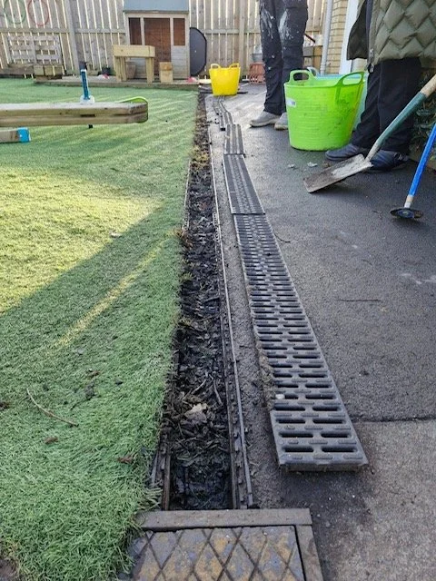 People working on a landscaping project, installing a drainage system along the edge of a lawn and concrete walkway, with tools and buckets nearby.