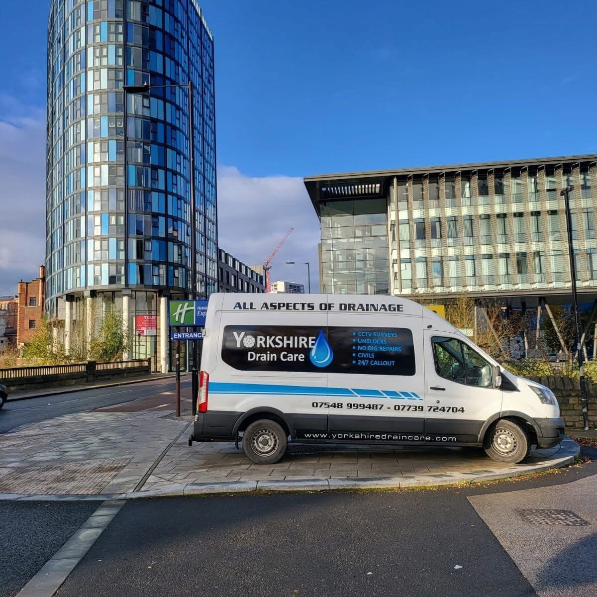 A white Yorkshiredraincare van parked on a paved sidewalk in front of modern glass buildings on a sunny day with a blue sky and some clouds.