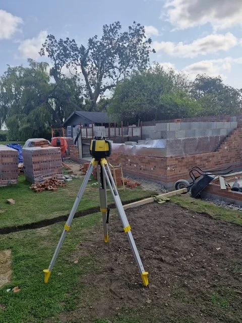 Construction site with a leveling instrument on tripod, bricks, and partially built brick wall in backyard with trees and shed in background.