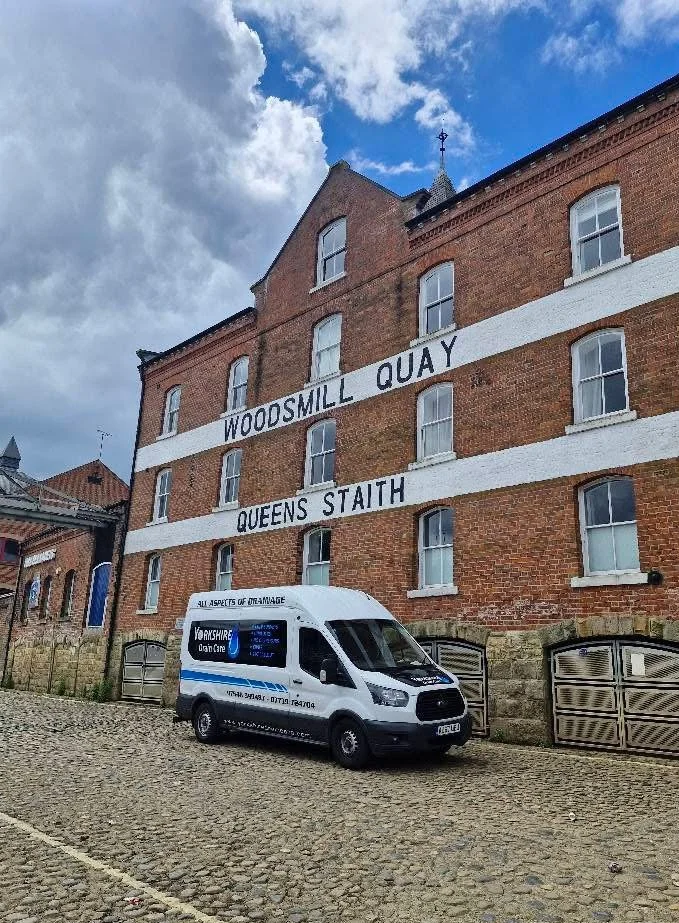 A red brick building with white window frames and large painted letters on the side saying 'WOODSILL QUAY QUEENS STAITH'. In front of the building, a white van with blue signage is parked on a cobblestone street under a partly cloudy sky.