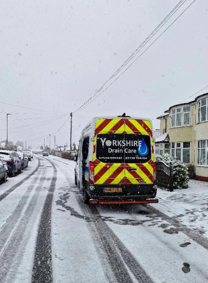A Yorkshires Drain Care service van on a snowy residential street, with snow-covered houses and parked cars, and tire tracks in the snow.