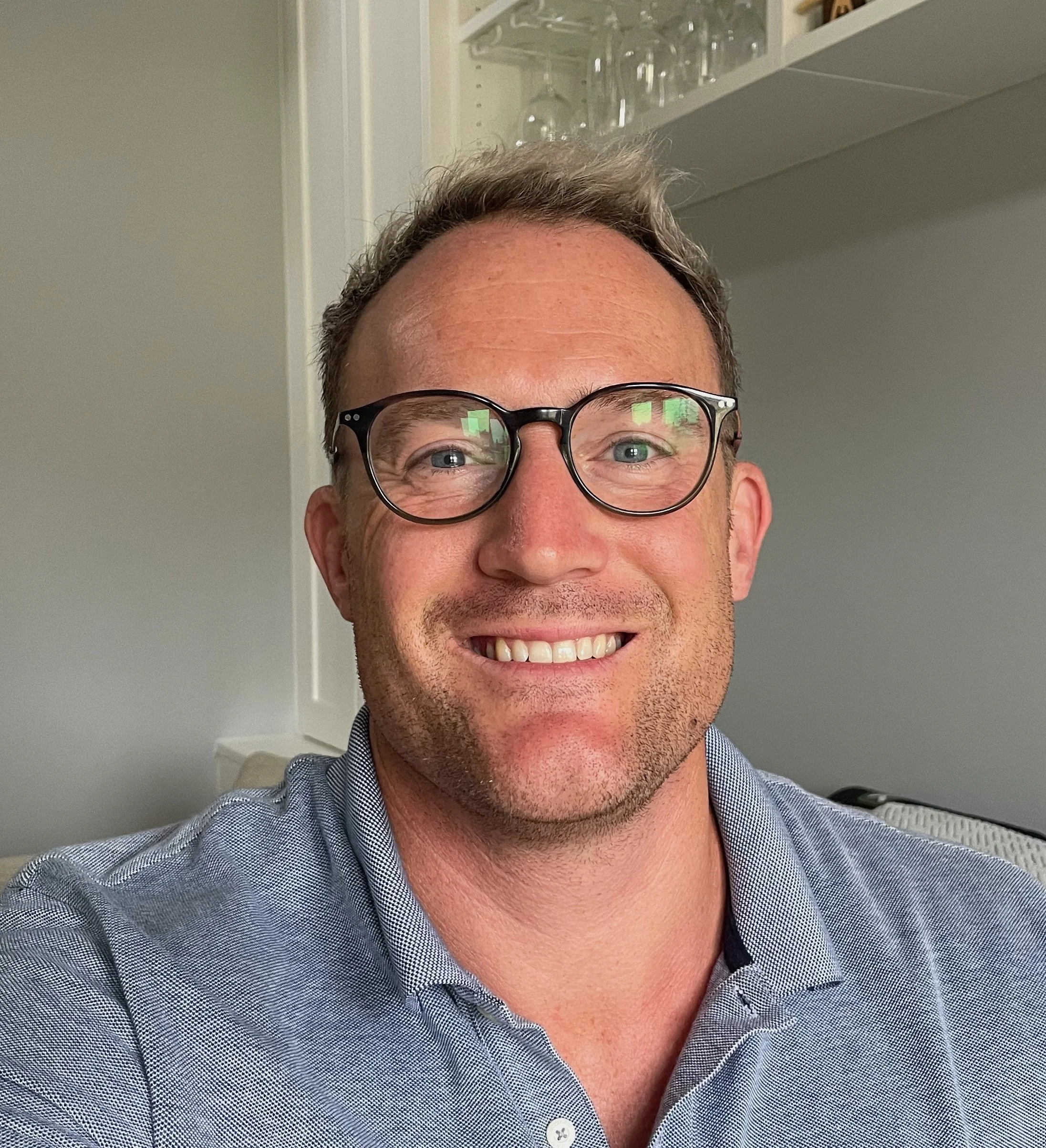 A close-up photo of a man with glasses smiling, showing his teeth. He is wearing a checkered shirt and has short hair. The background includes a white wall and a shelf with glassware.