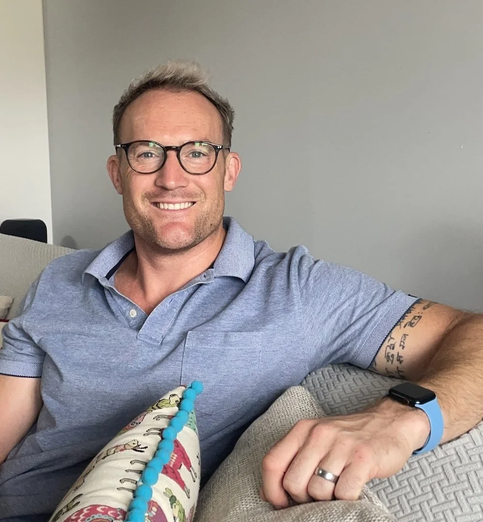A smiling man with glasses, wearing a gray polo shirt, sitting on a beige couch in a living room.