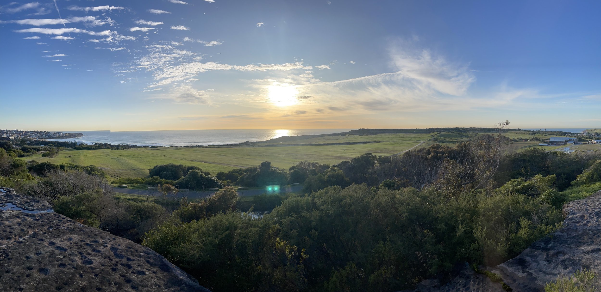 A scenic view of a coastal landscape during sunset, with green fields, trees, and a distant shoreline with buildings, under a partly cloudy sky.