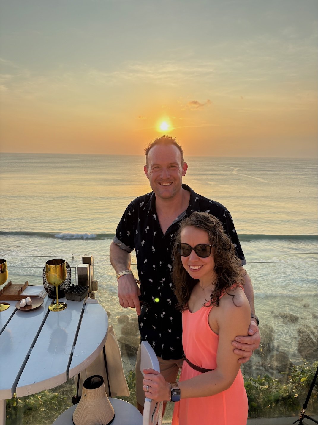 A smiling man and woman stand together on a beach at sunset, with the ocean and sky in the background. The woman wears sunglasses and a bright pink dress, while the man wears a black shirt with a white pattern. They are near a table with gold-colored cups and some objects, and appear to be enjoying a relaxing moment by the water.