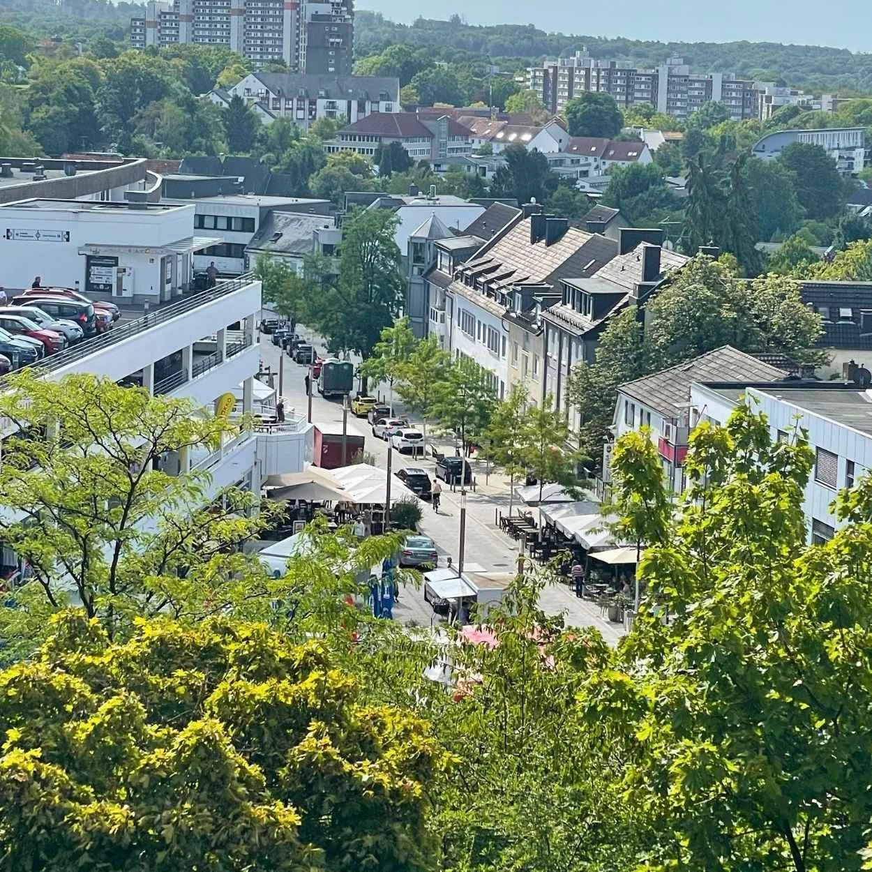Schloßstraße von oben: Ein Stadtbild mit mehrstöckigen Gebäuden, Bäumen und einer Straße mit parkenden Autos und Cafés im Vordergrund.