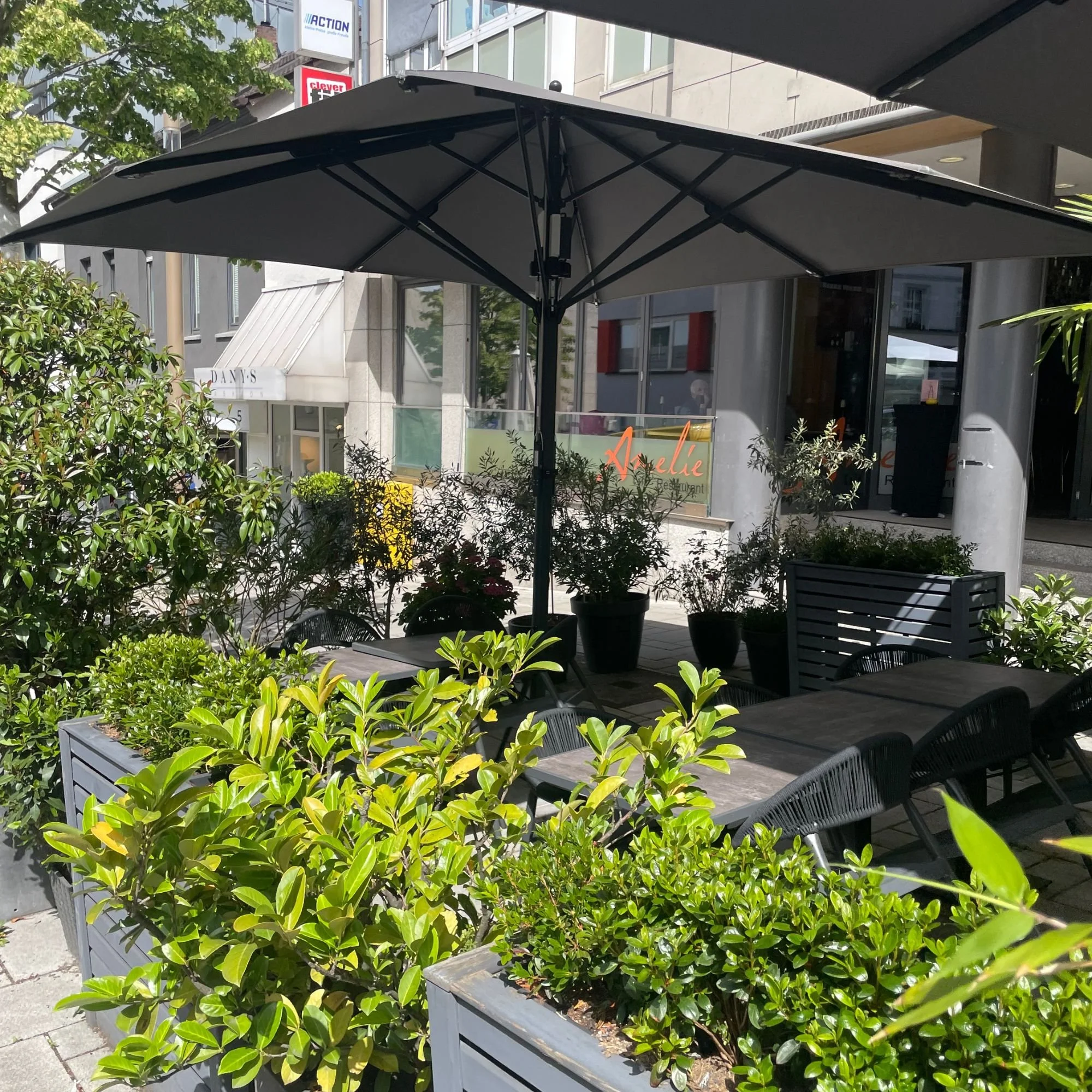 Schloßstarße Bensberg: Ein schwarzer Sonnenschirm auf einer Terrasse, umgeben von grünen Büschen und Möbeln, mit einem Geschäftsgebäude im Hintergrund.