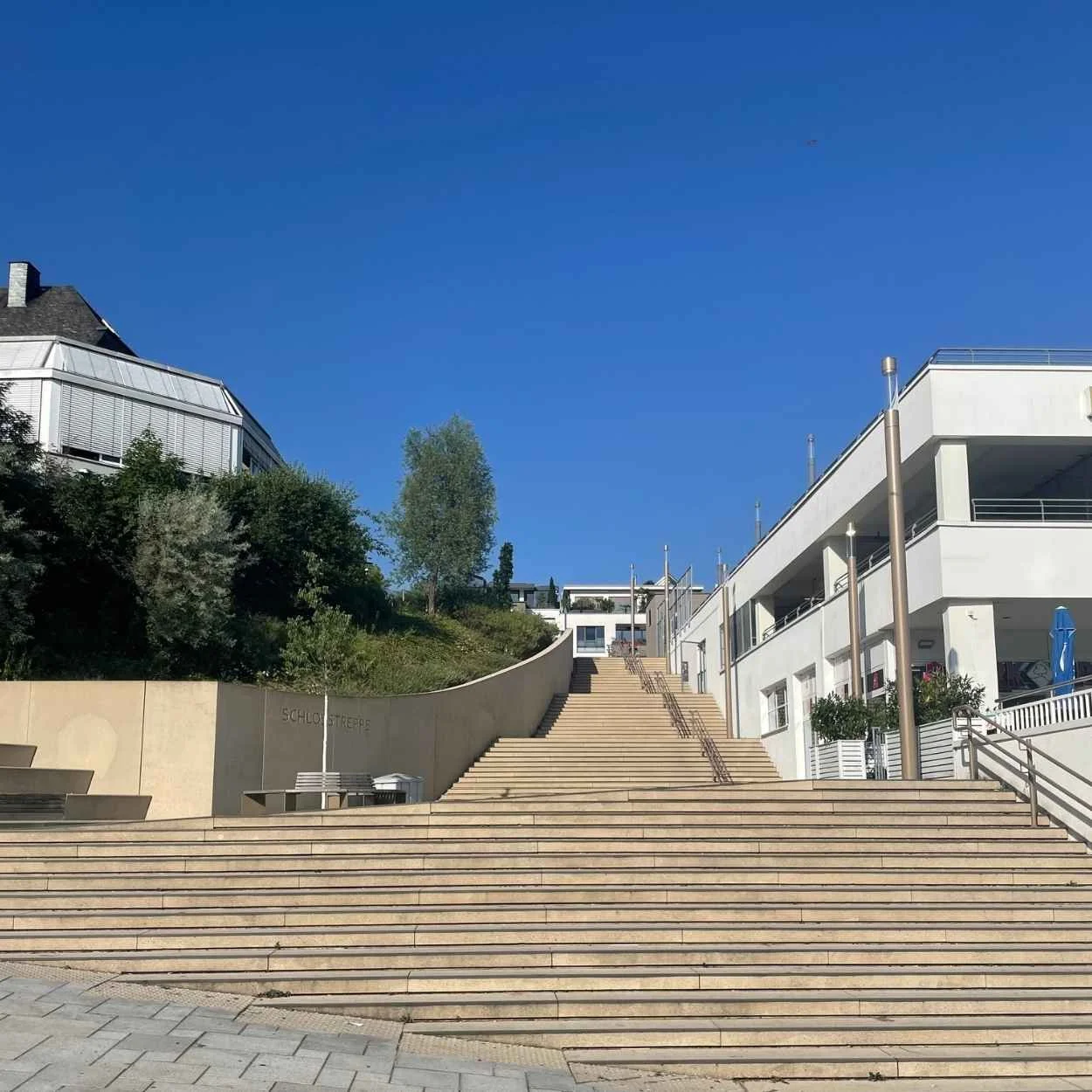 Schloßtreppe Bensberg: Stufen und Treppe führen zu einem modernen Einkaufscenter mit weißen Fassaden, umgeben von Bäumen und einem blauen Himmel.