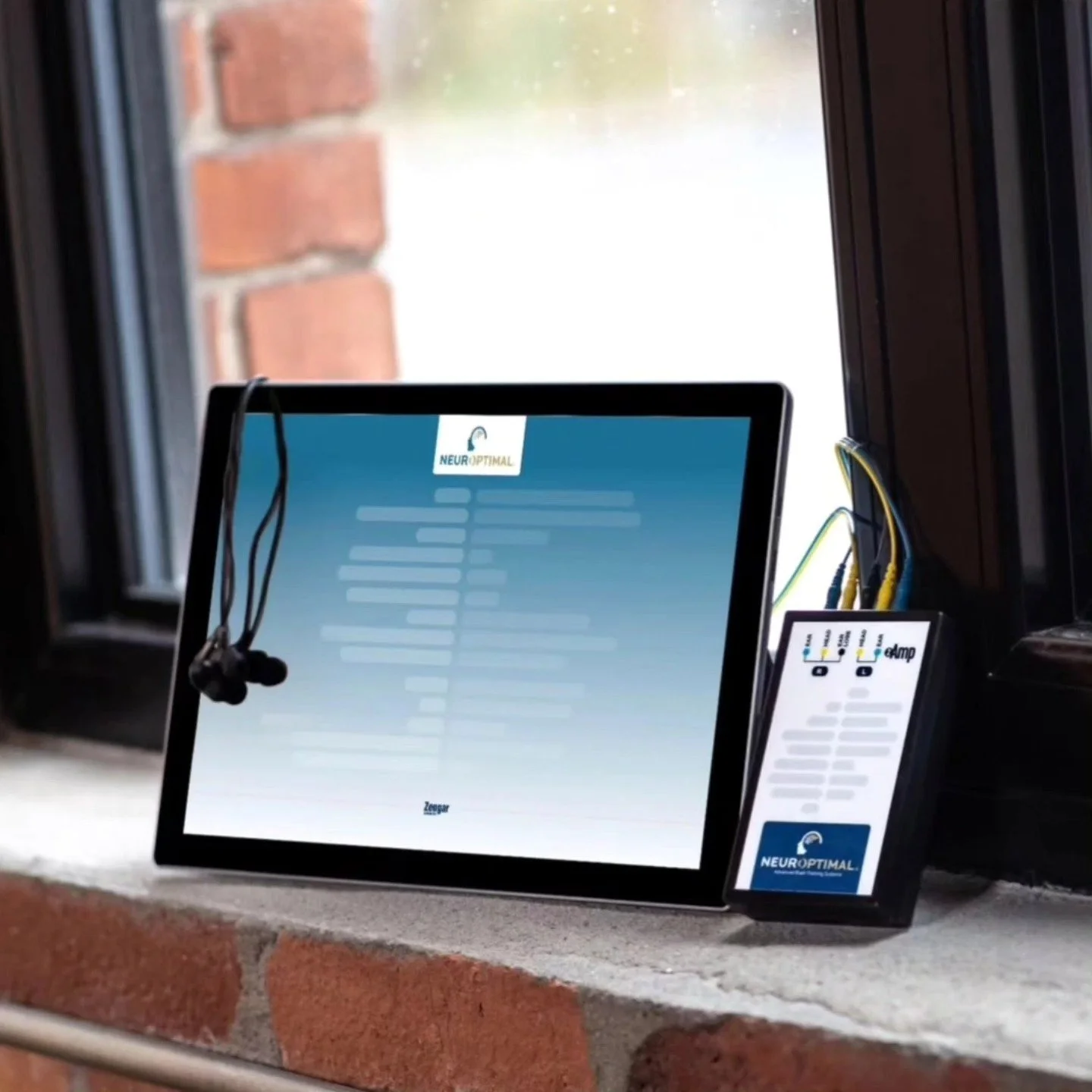 A tablet and smartphone displaying the NeuroOptimal website and logo, with a pair of black earbuds hanging near the tablet, set on a brick ledge next to a window.