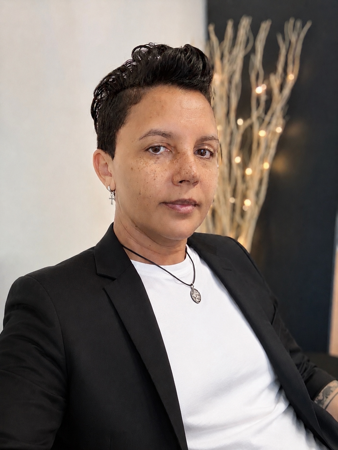 A woman with short dark hair, light skin with freckles, wearing a black blazer over a white shirt, silver cross earring, necklace with a tree of life pendant, sitting in front of a decorative branch with lights.